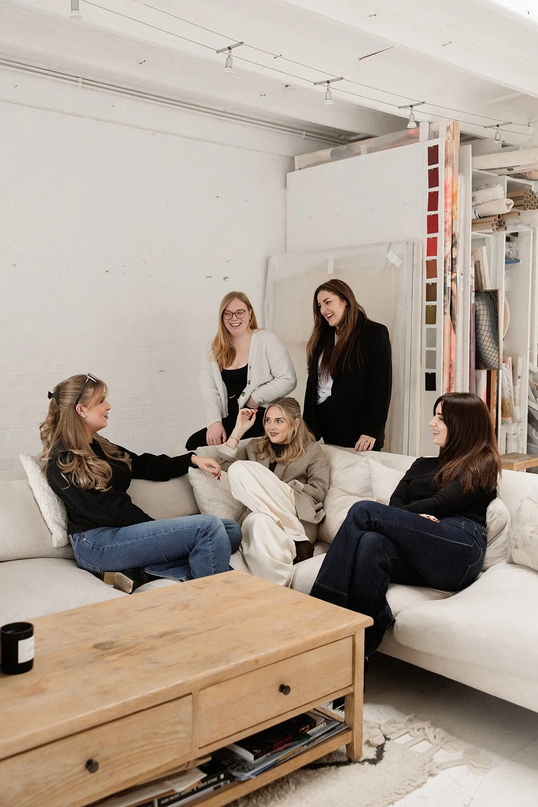 Group of five women smiling and chatting in a cozy living room, sitting on and standing around a white sectional sofa, with a wooden coffee table in front, and a white brick wall behind them.
