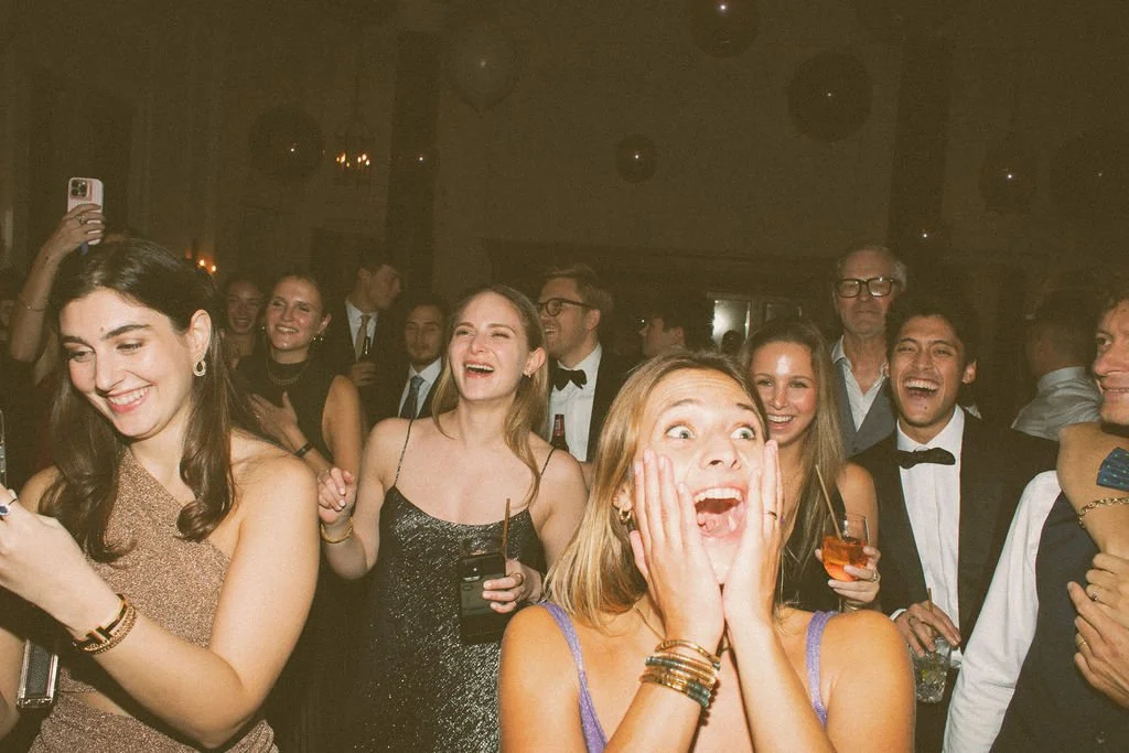 Group of people in formal attire at a celebration or party, some are laughing and smiling, with one woman in the foreground reacting with surprise or excitement.