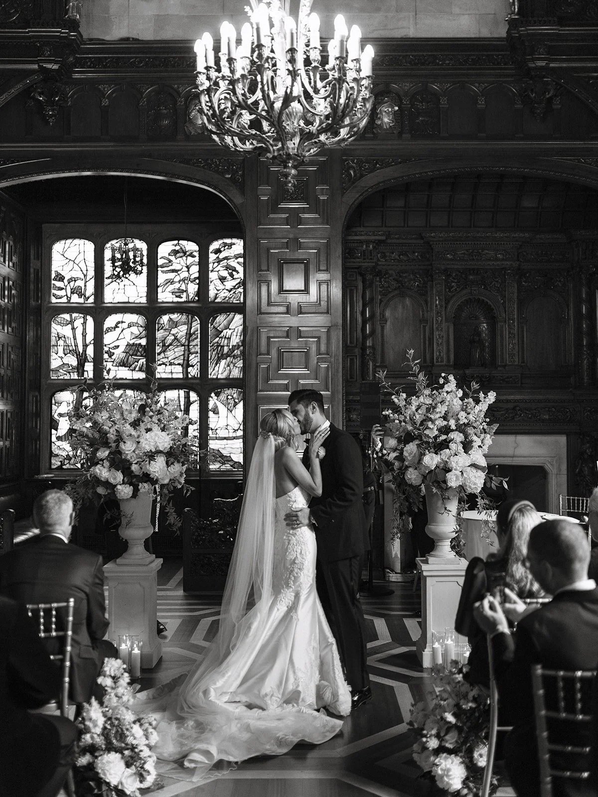 A black-and-white photo of a wedding ceremony in a grand, ornate room. The bride and groom are holding each other and about to kiss, standing in front of stained glass windows and floral arrangements. Guests are seated around them, watching the moment.