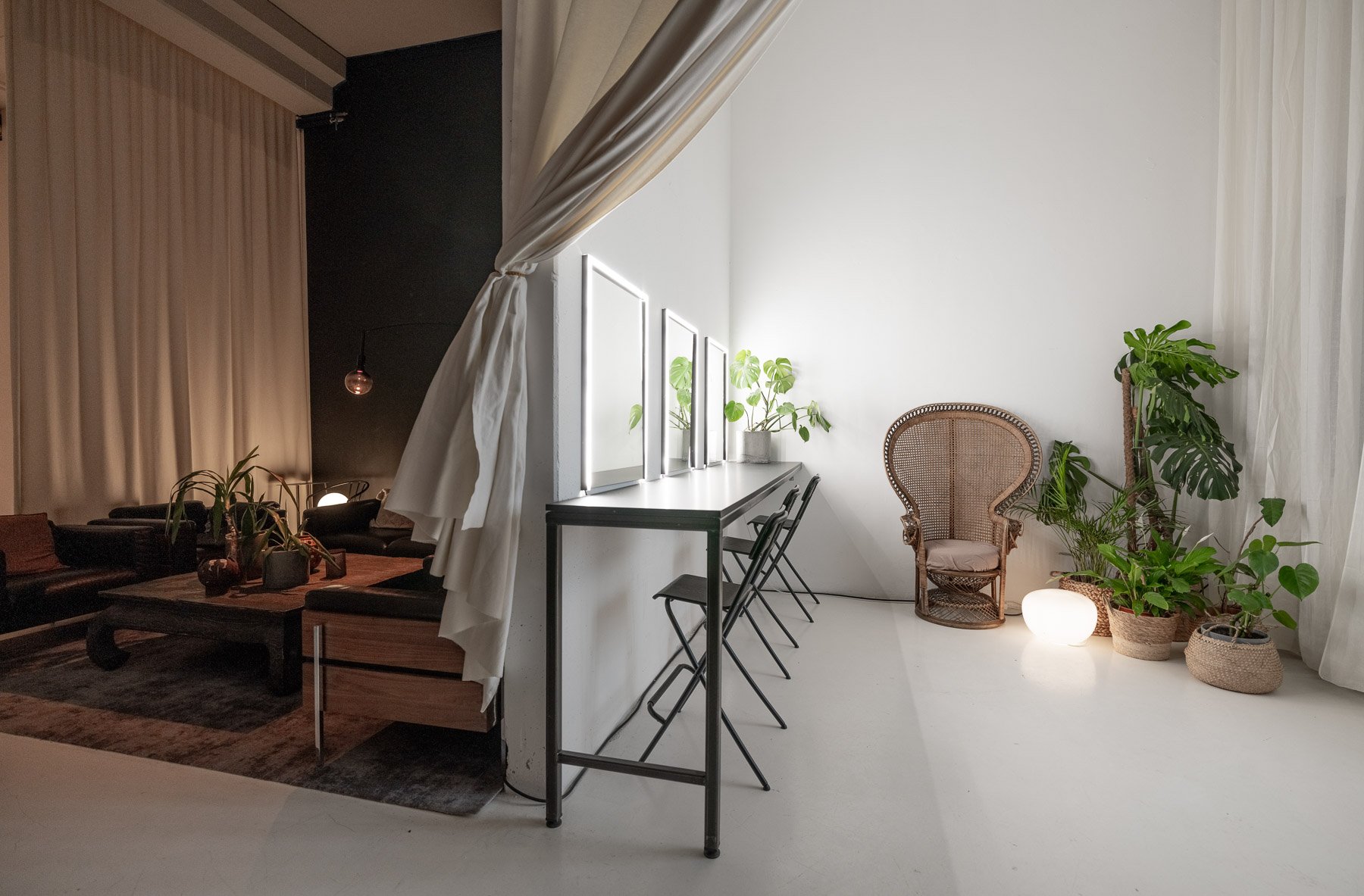 Interior of a modern, minimalist room with a white wall and a high ceiling. On the right, there are several potted plants, a wicker armchair, and a glowing small lamp. On the left, there is a white countertop with black chairs arranged along it, and 