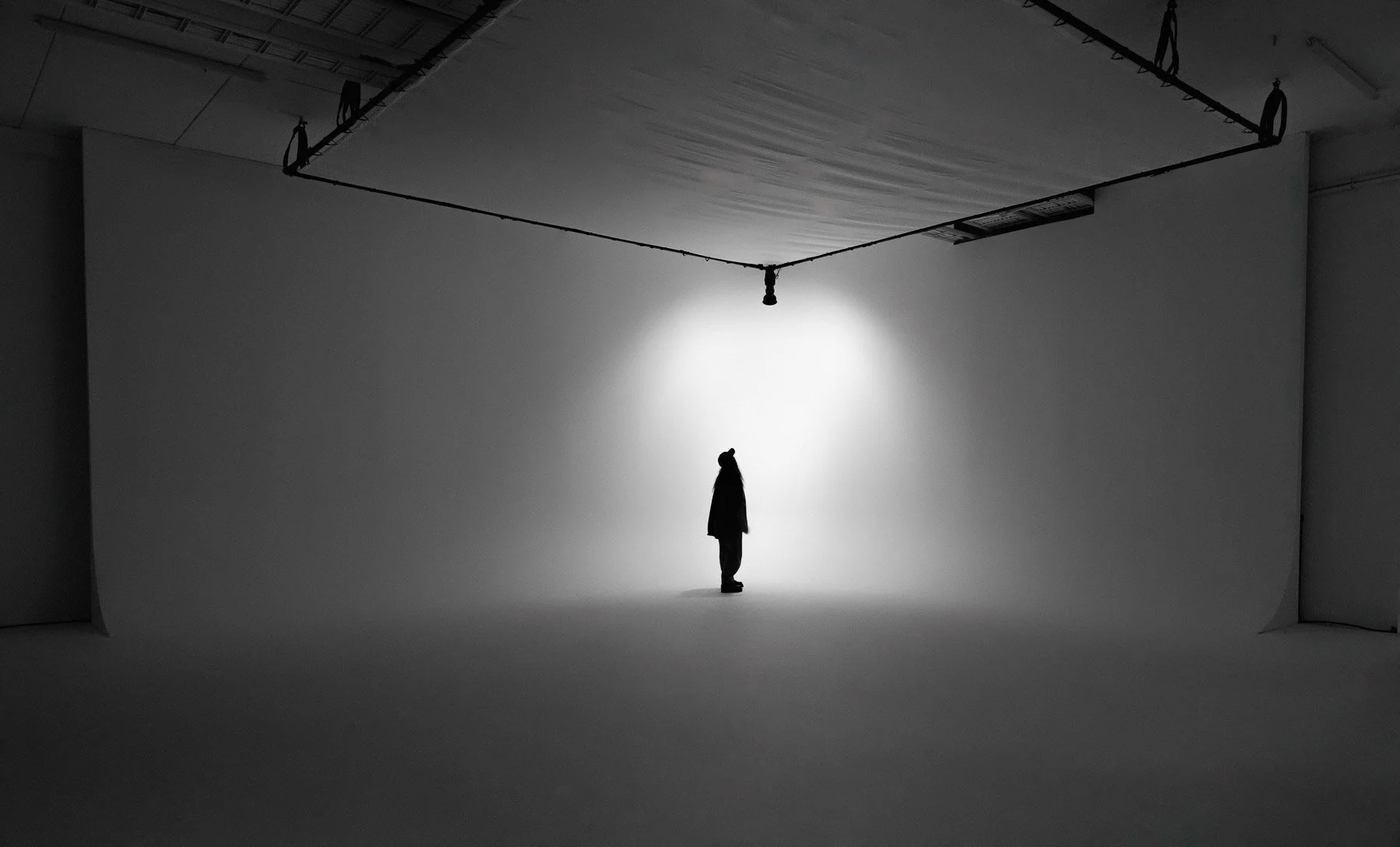 A person standing in a photo studio with plain white walls and a large overhead light, silhouette style in black and white.