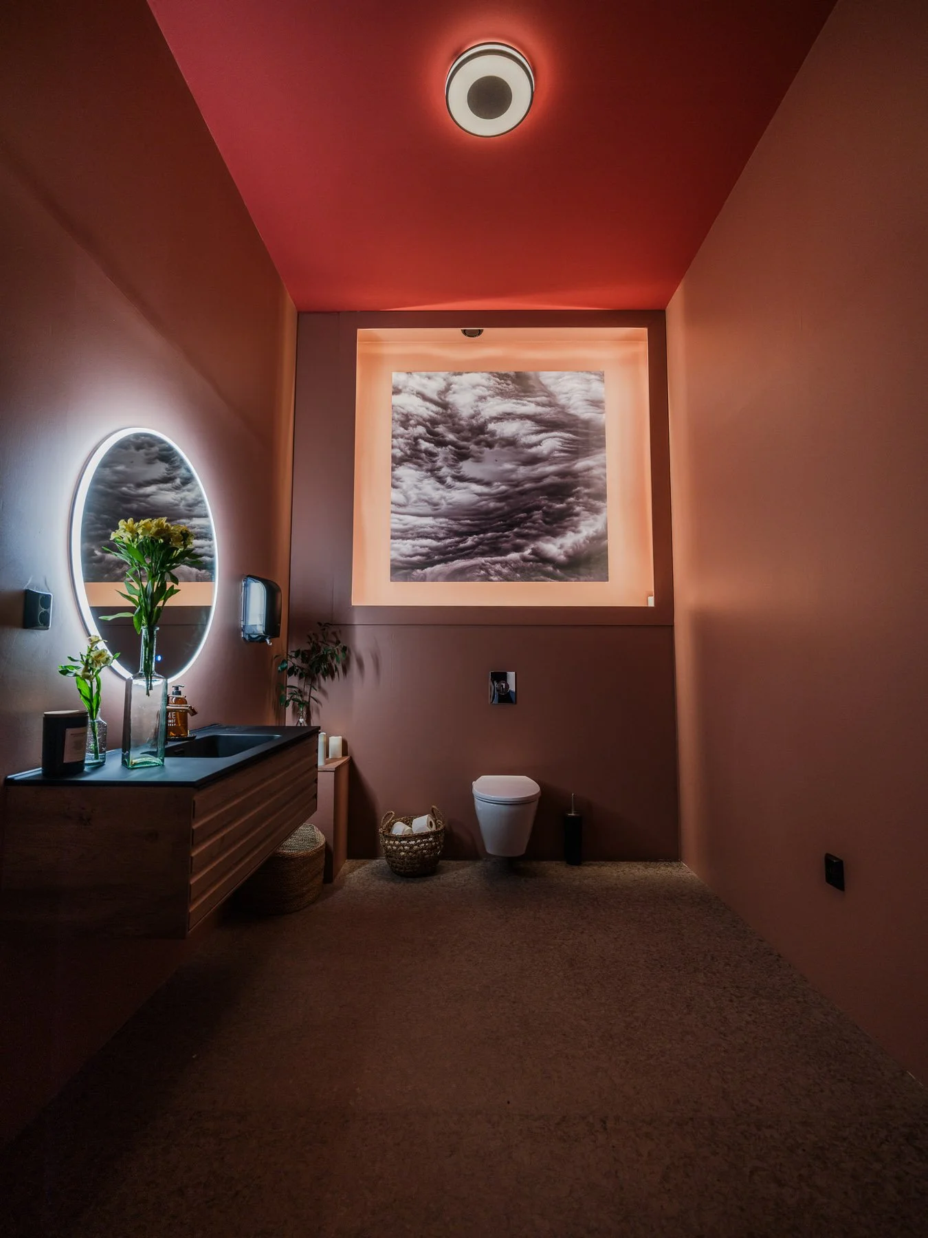 A modern bathroom with pink walls and ceiling, illuminated by soft lighting. There is a round illuminated mirror above a wooden vanity with flowers and toiletries. A toilet is situated below a large window with a view of cloudy sky.