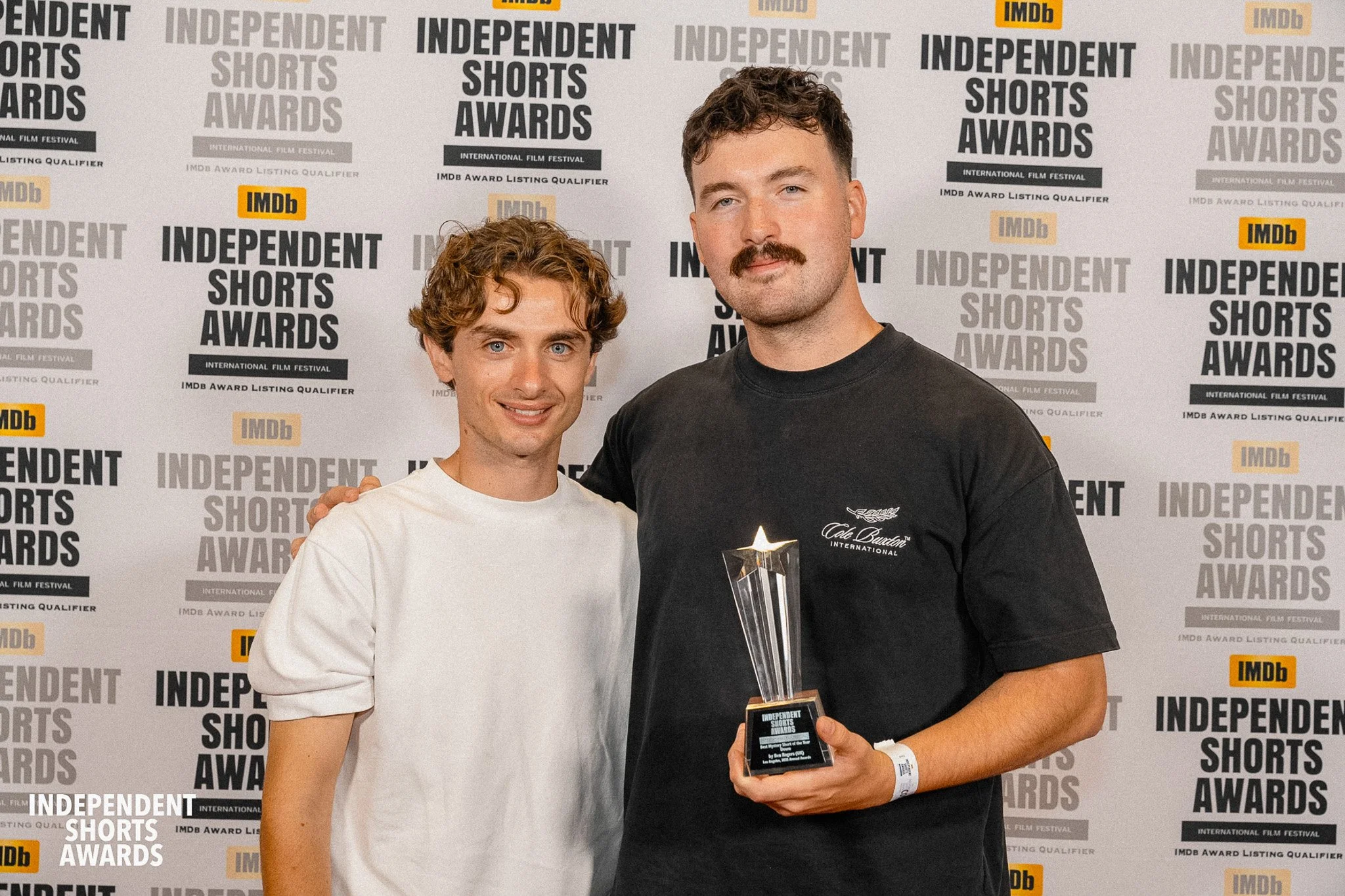 Two men posing together at an awards event with a backdrop displaying 'Independent Shorts Awards' and 'IMDb' logos; one man is holding a trophy.