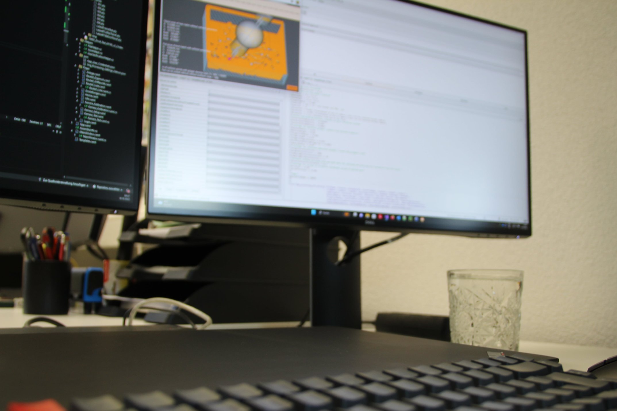 Ein Arbeitsplatz mit zwei Monitoren, einem Keyboard, Stifte in einem Behälter und einem Wasserglas auf dem Tisch. Die Monitore zeigen Programmier- oder Entwicklungstools.