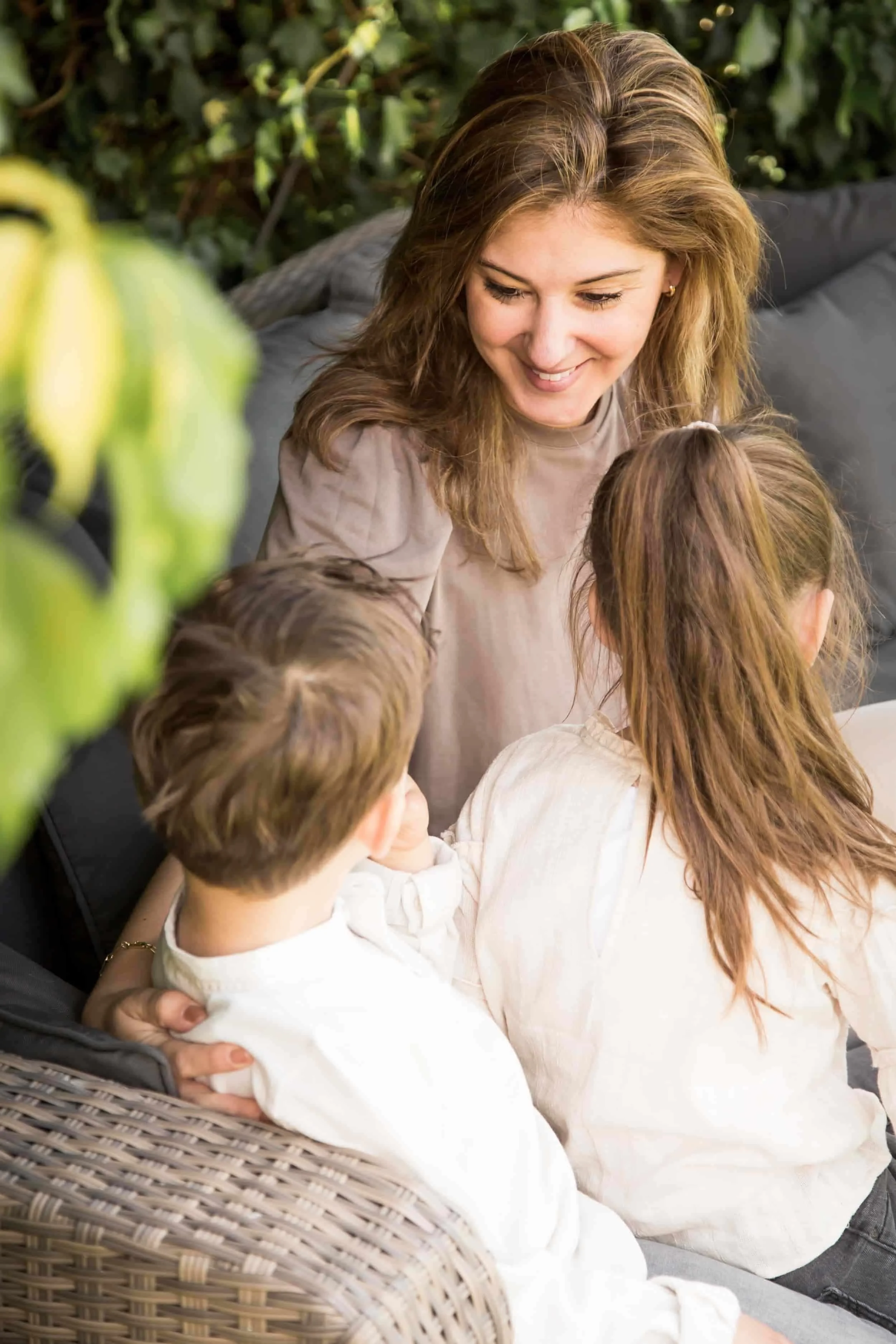 Vrouw strijkt met vier jonge kinderen buiten in een tuin, terwijl ze allemaal op een bankje zitten. Er is groen en bladeren op de achtergrond.