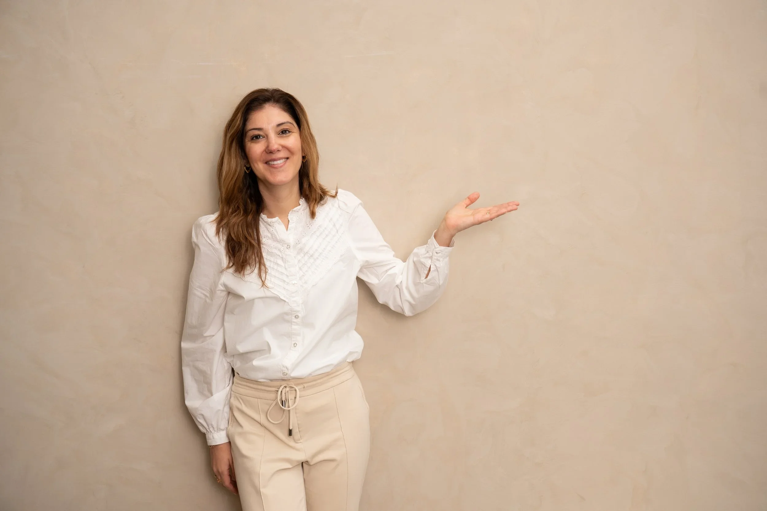 Vrouw met lang, kastanjebruin haar, glimlachend en wijzend naar rechts, staand tegen een beige muur, gekleed in een witte blouse en beige joggingsbroek.