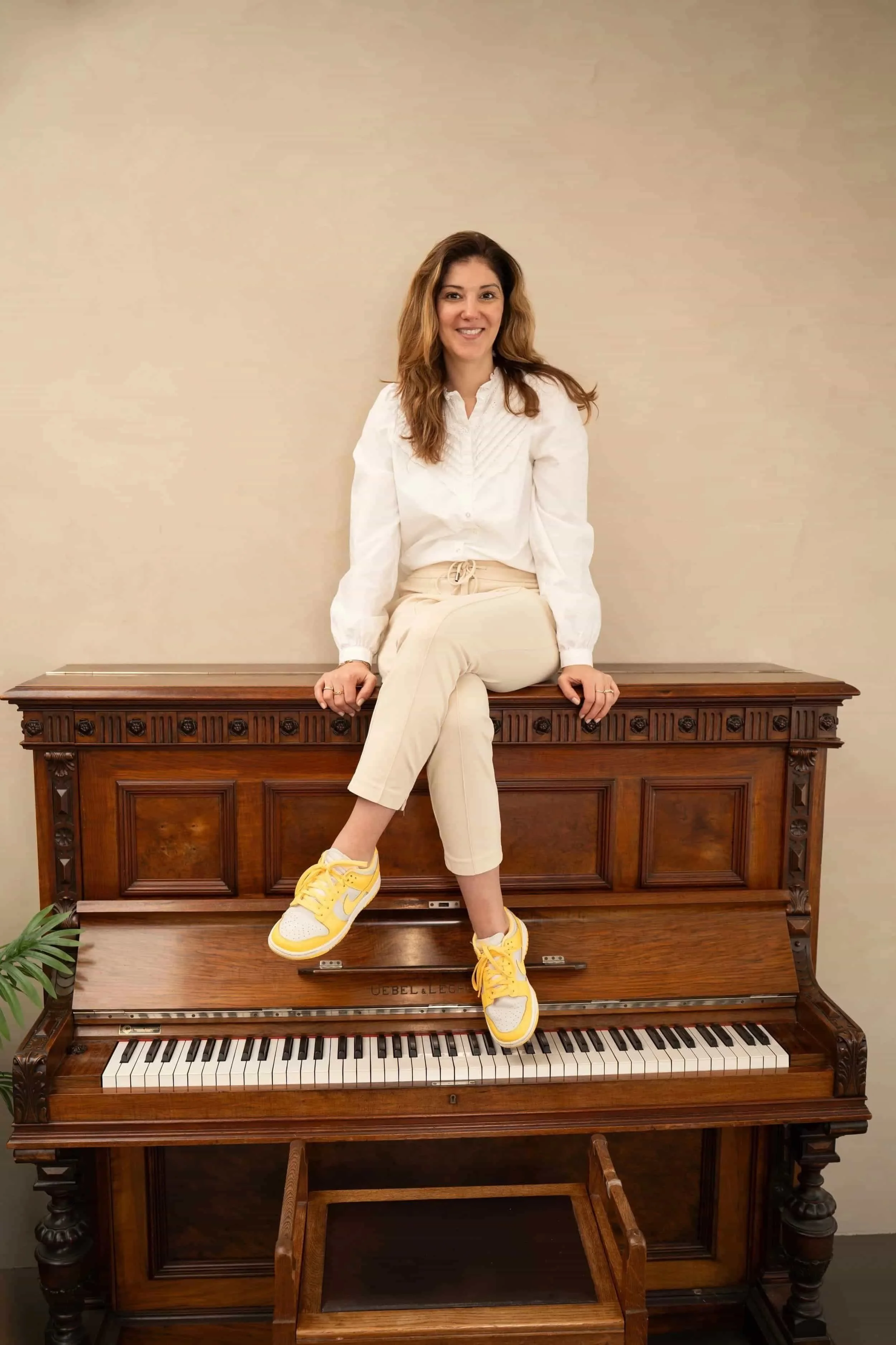 Vrouw zit op een oude houten piano met het deksel open, kleed zich casual in een witte blouse en beige broek, en draagt gele sneakers, in een kamer met een effen lichte muur.