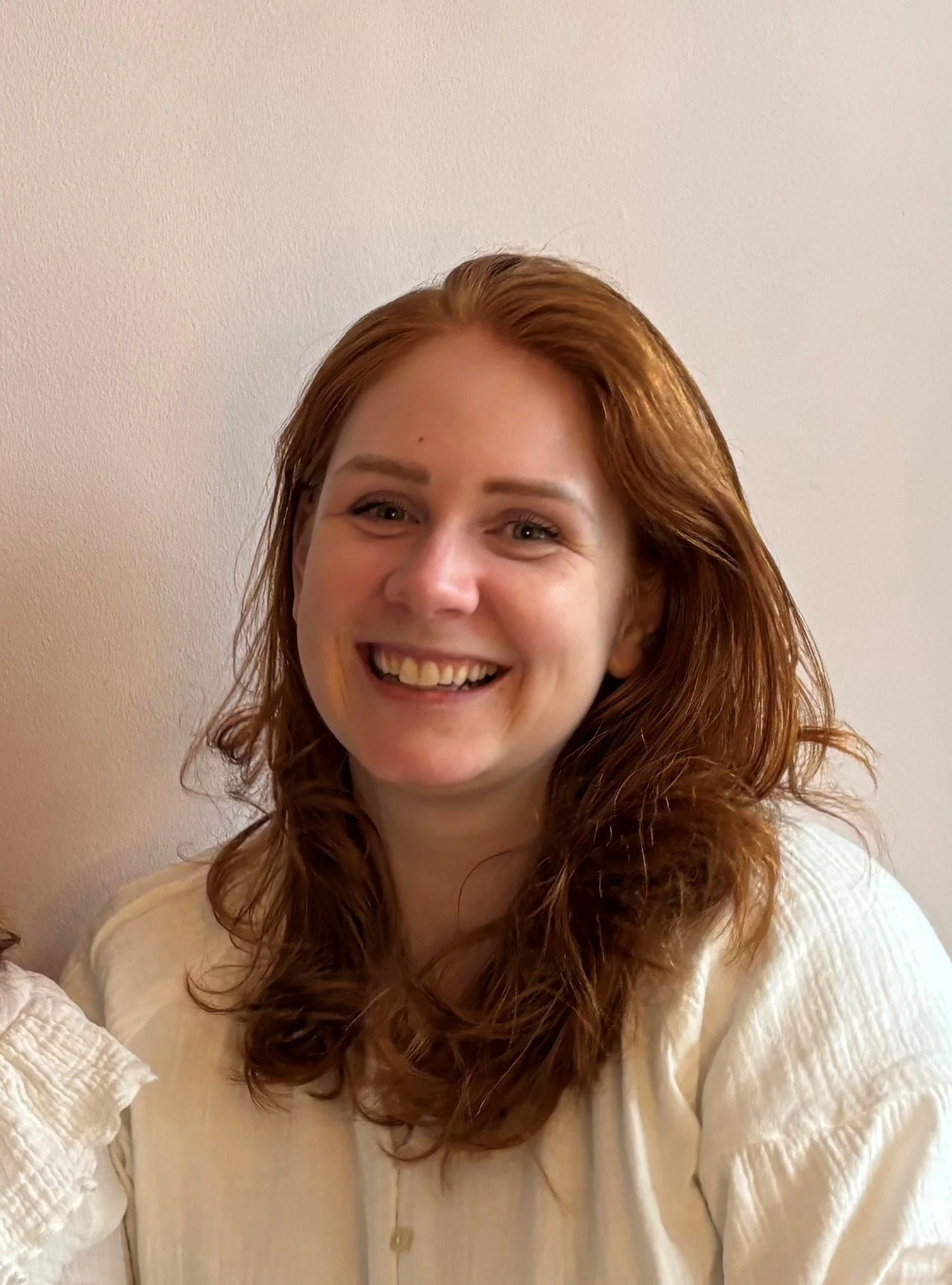 Een lachende vrouw met rood haar, gekleed in een witte blouse, voor een lichte achtergrond.