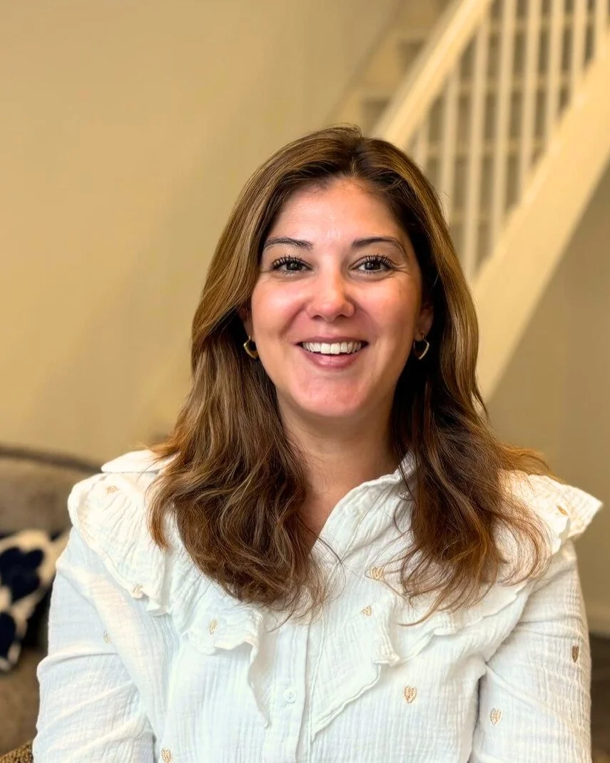 Een lachende vrouw met bruin haar en een witte blouse, zittend in een huis.