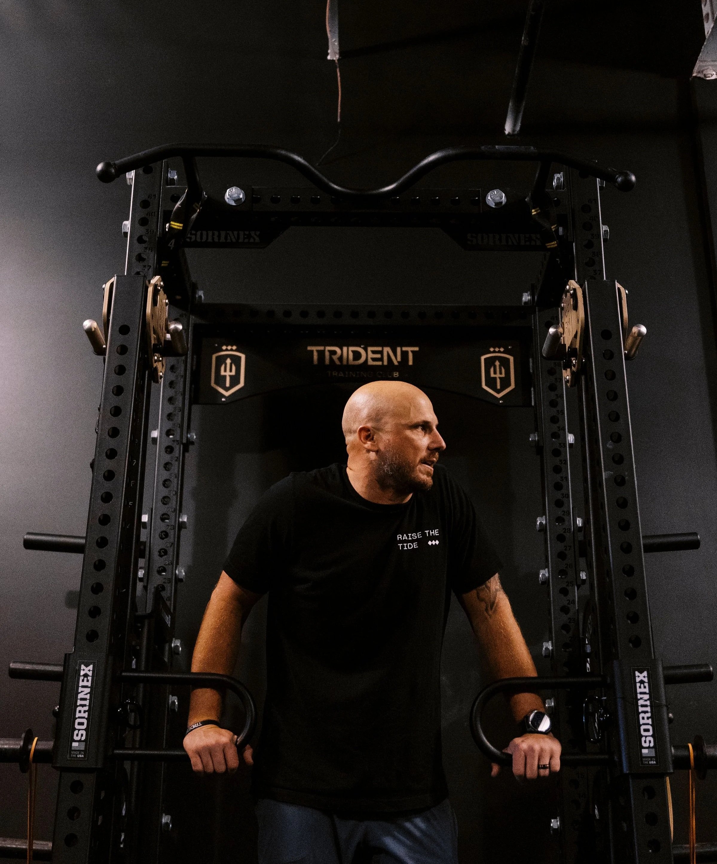 A man with a shaved head and beard, wearing a black t-shirt, training with a harness or chest supporter in a gym.