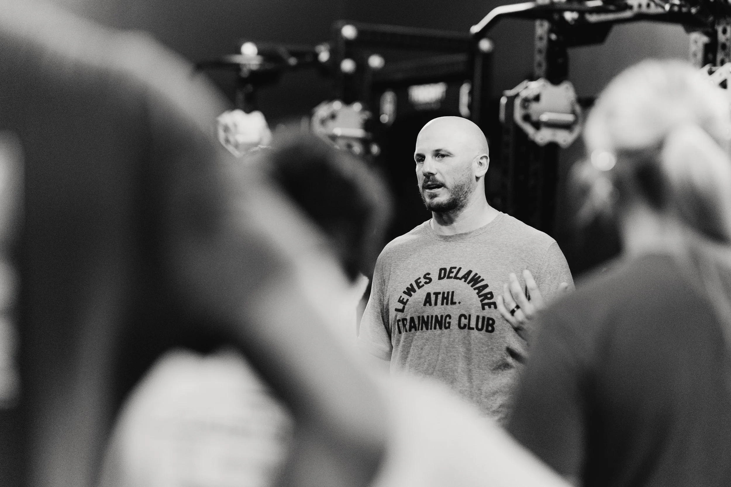 A male coach speaking to a group of people in a gym or fitness training class, blurred foreground, in black and white.
