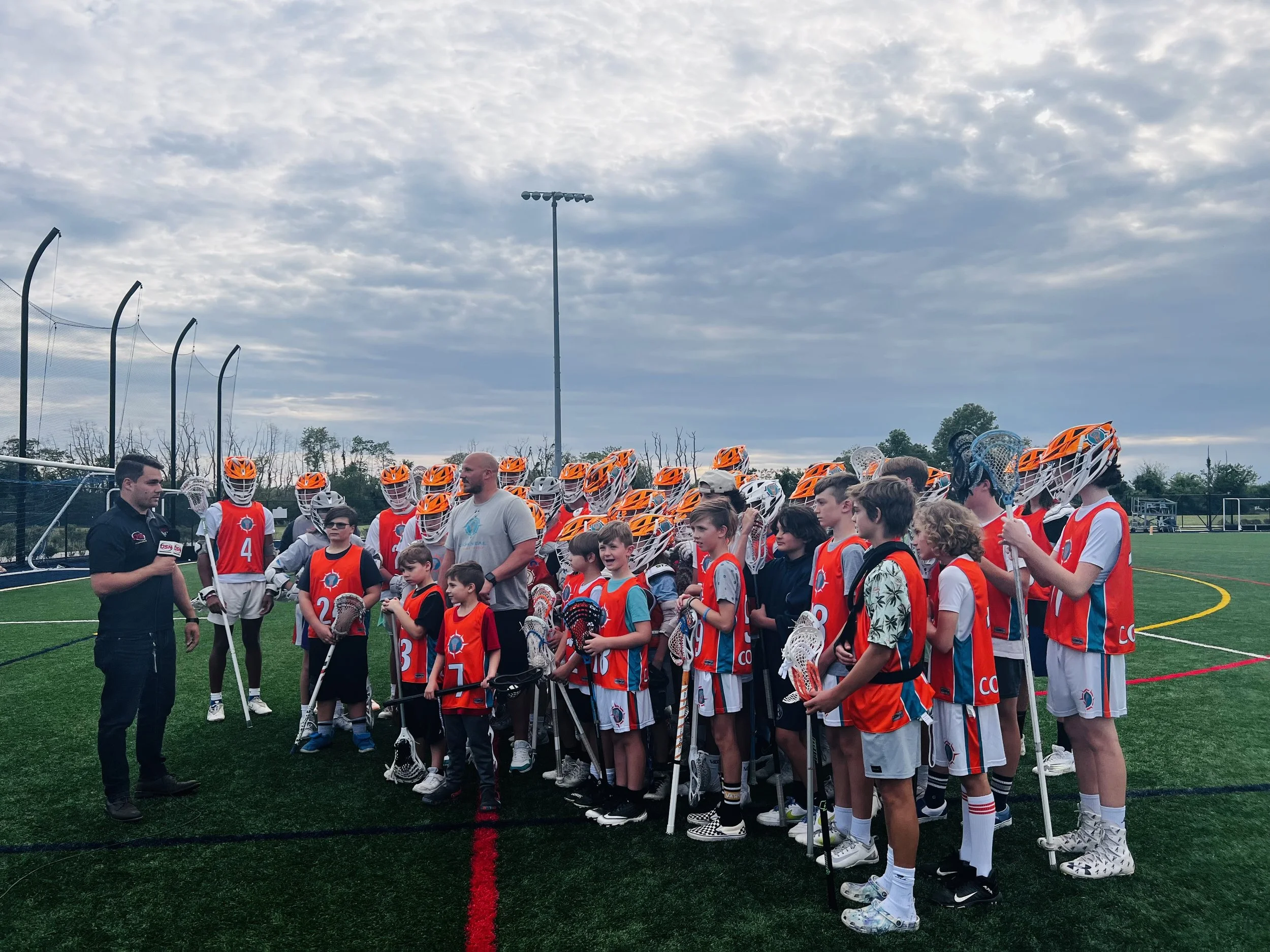 Lacrosse team with young players and coaches on a sports field during a team gathering, with a coach speaking to the group, under a cloudy sky.