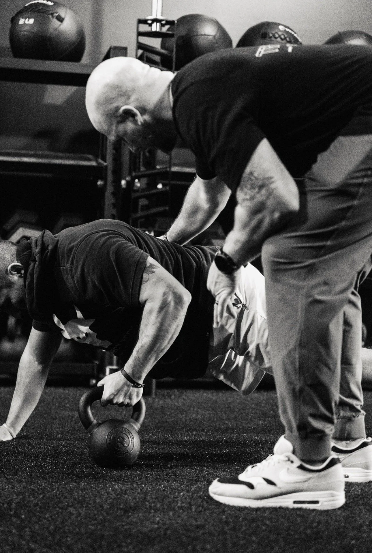 A man doing a push-up with a kettlebell on the ground in a gym, with a trainer assisting him. Gym equipment, including medicine balls, is visible in the background.