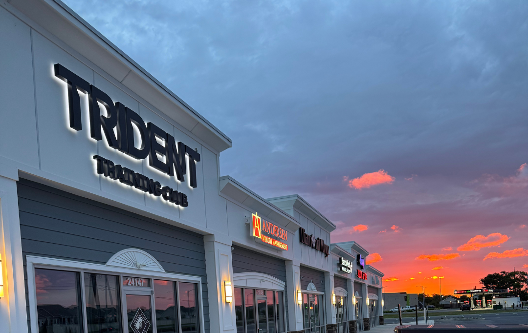 Shopping plaza with retail stores including Trident Trading Co, Andersen Wealth Management, and others at sunset, with colorful clouds and a parking lot in the background.