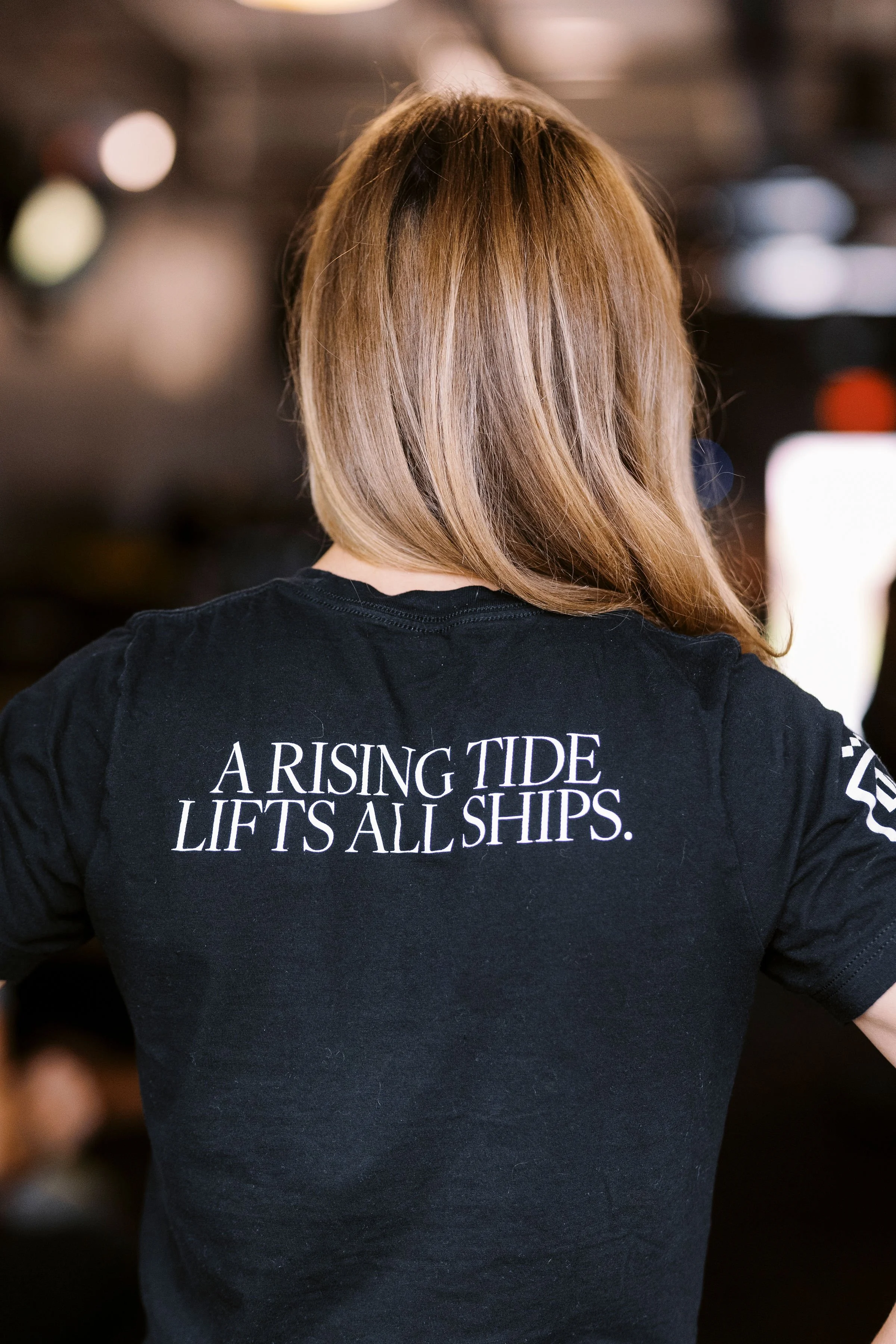 Back of a person wearing a black T-shirt with white text that reads, "A RISING TIDE LIFTS ALL SHIPS". The person has light brown hair and is indoors with a blurred background.