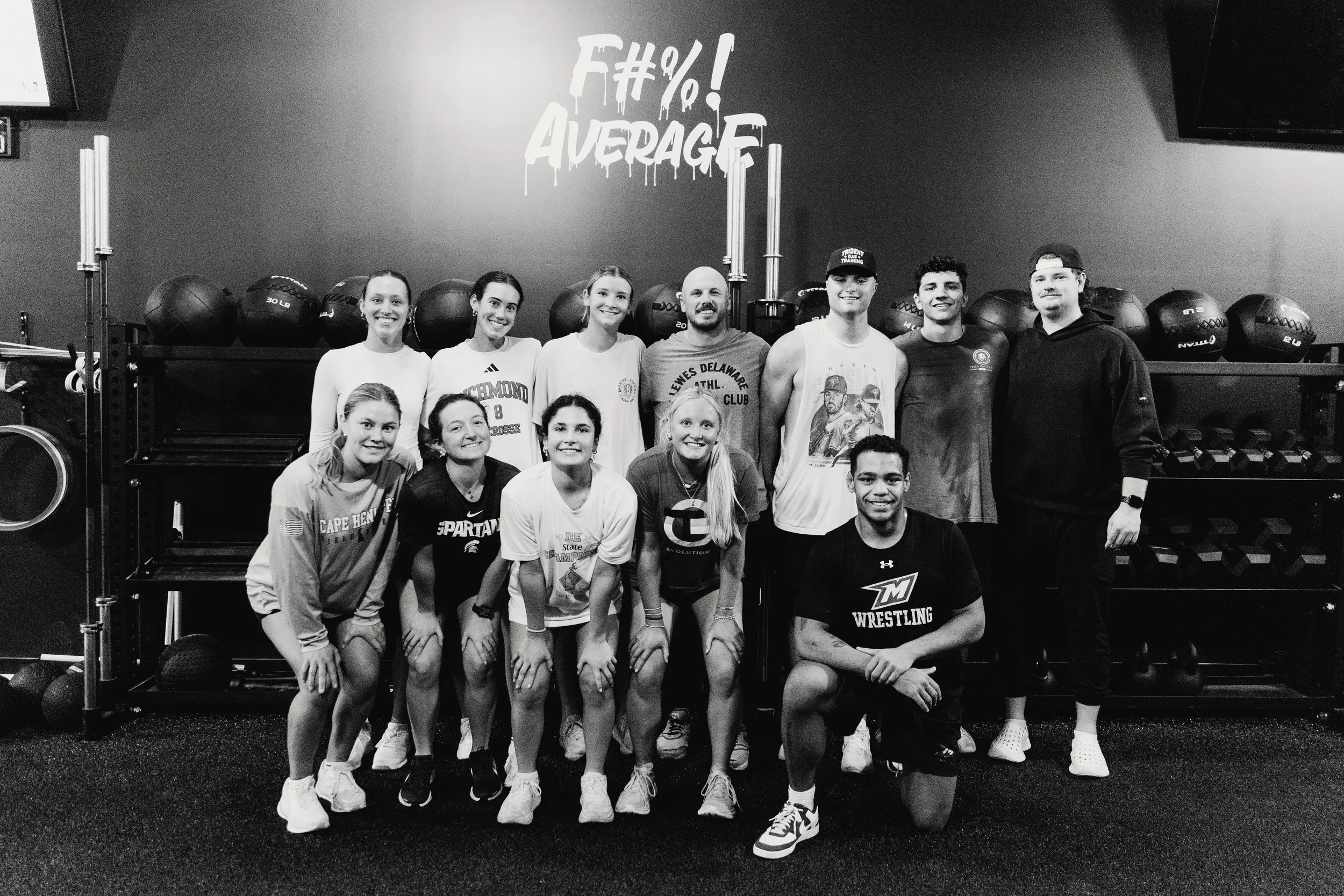 Group of eleven young adults in a gym, some wearing athletic shirts and shorts, posing for a photo with exercise equipment in the background and a wall sign that says 'F#%!. AVERAGE'.