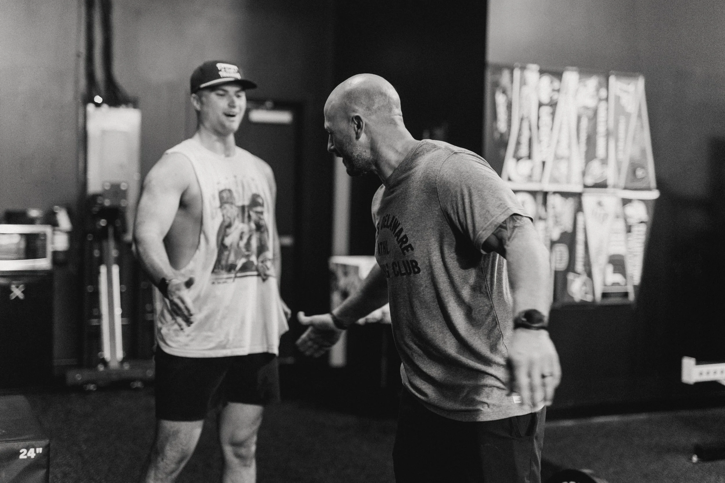 Two men in a gym, one with his shirt off, and the other in a t-shirt gesturing with his hand, engaging in a conversation or coaching session.