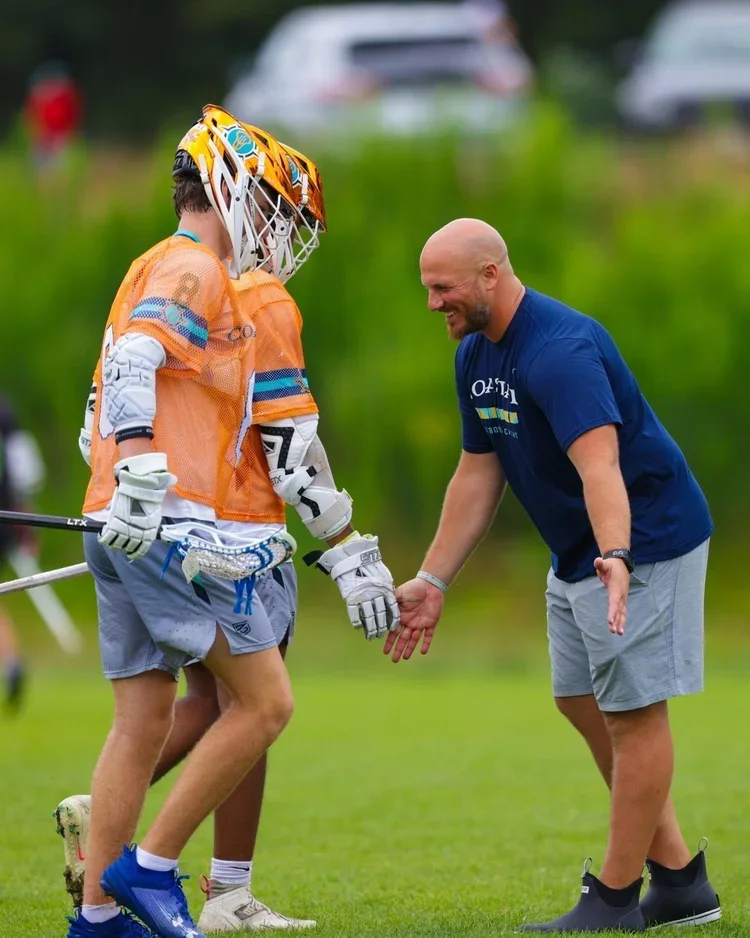 A man in a navy t-shirt and gray shorts smiling as he shakes hands with a lacrosse player in an orange jersey and helmet on a grassy field.
