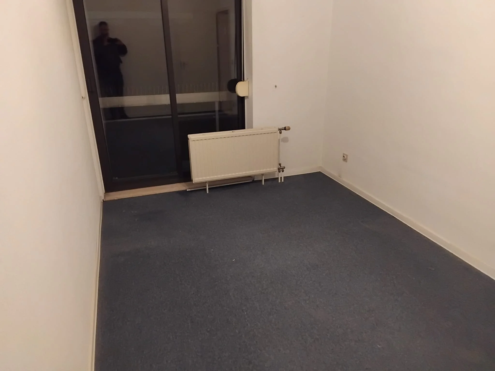 Une pièce vide avec des murs blancs, un radiateur blanc sous une fenêtre en verre, et un sol couvert de moquette foncée, avec une personne visible en réflexion dans la fenêtre.
