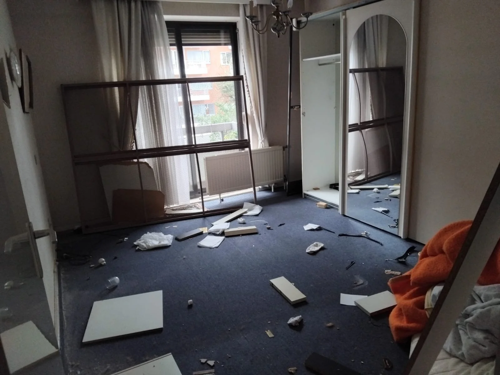 Une pièce en désordre avec des meubles brisés, des débris de verre, des morceaux de bois, et des vêtements, probablement suite à un acte de vandalisme ou une catastrophe.