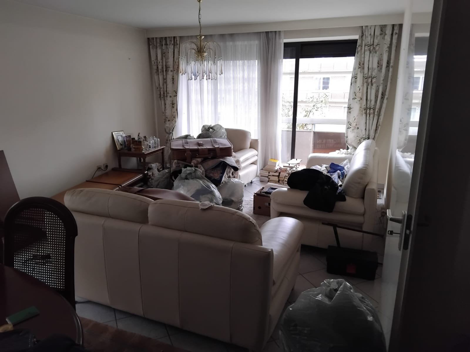 Salon encombré avec meubles en cuir blanc, des piles de livres, des sacs en plastique et des vêtements en désordre, linge sur les canapés, fenêtre avec rideaux et balcon en arrière-plan.