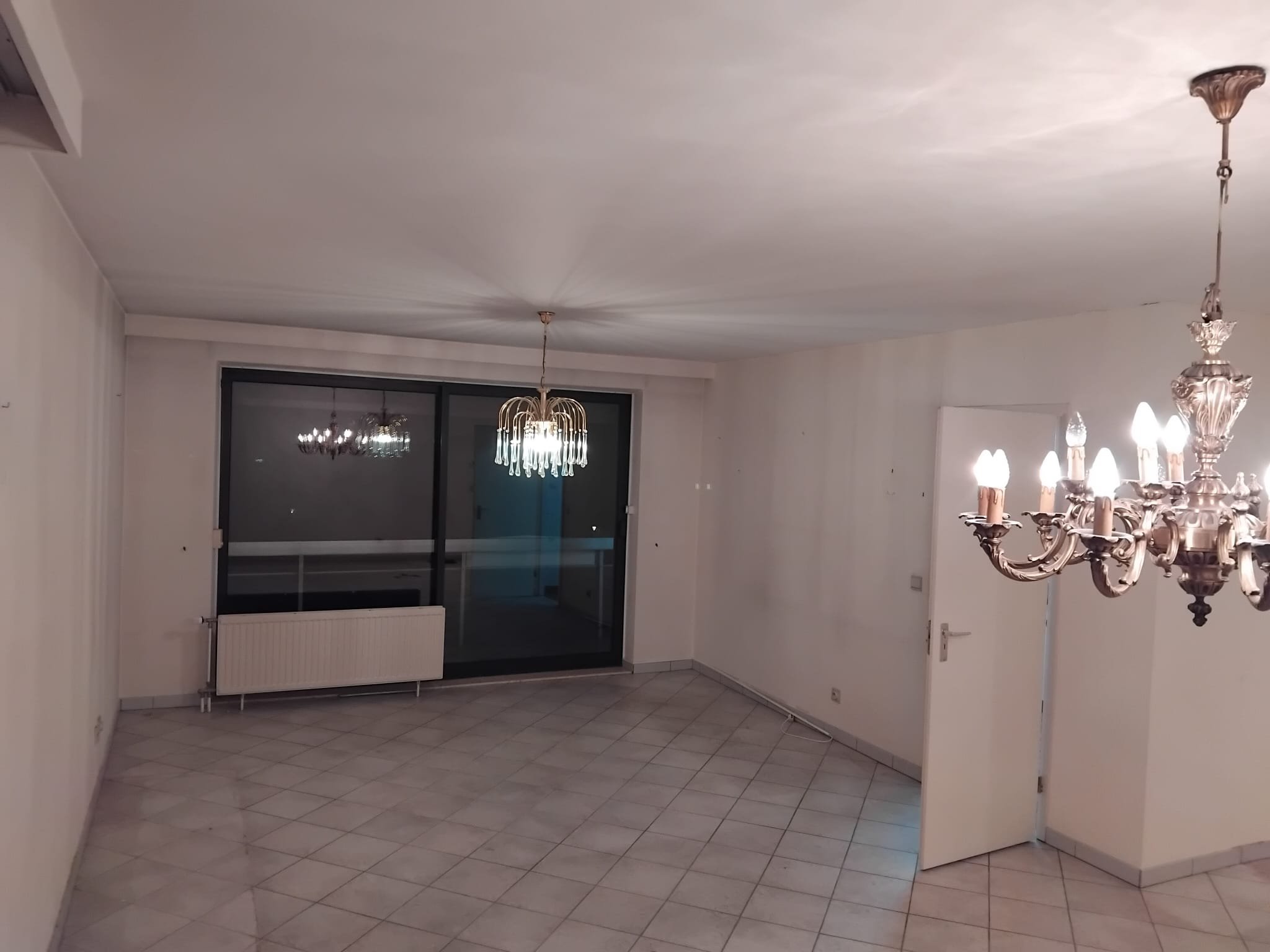 Salon vide avec deux lustres au plafond, un grand fenêtre avec volet roulant et un radiateur dessous. Le sol est carrelé et les murs sont peints en blanc.