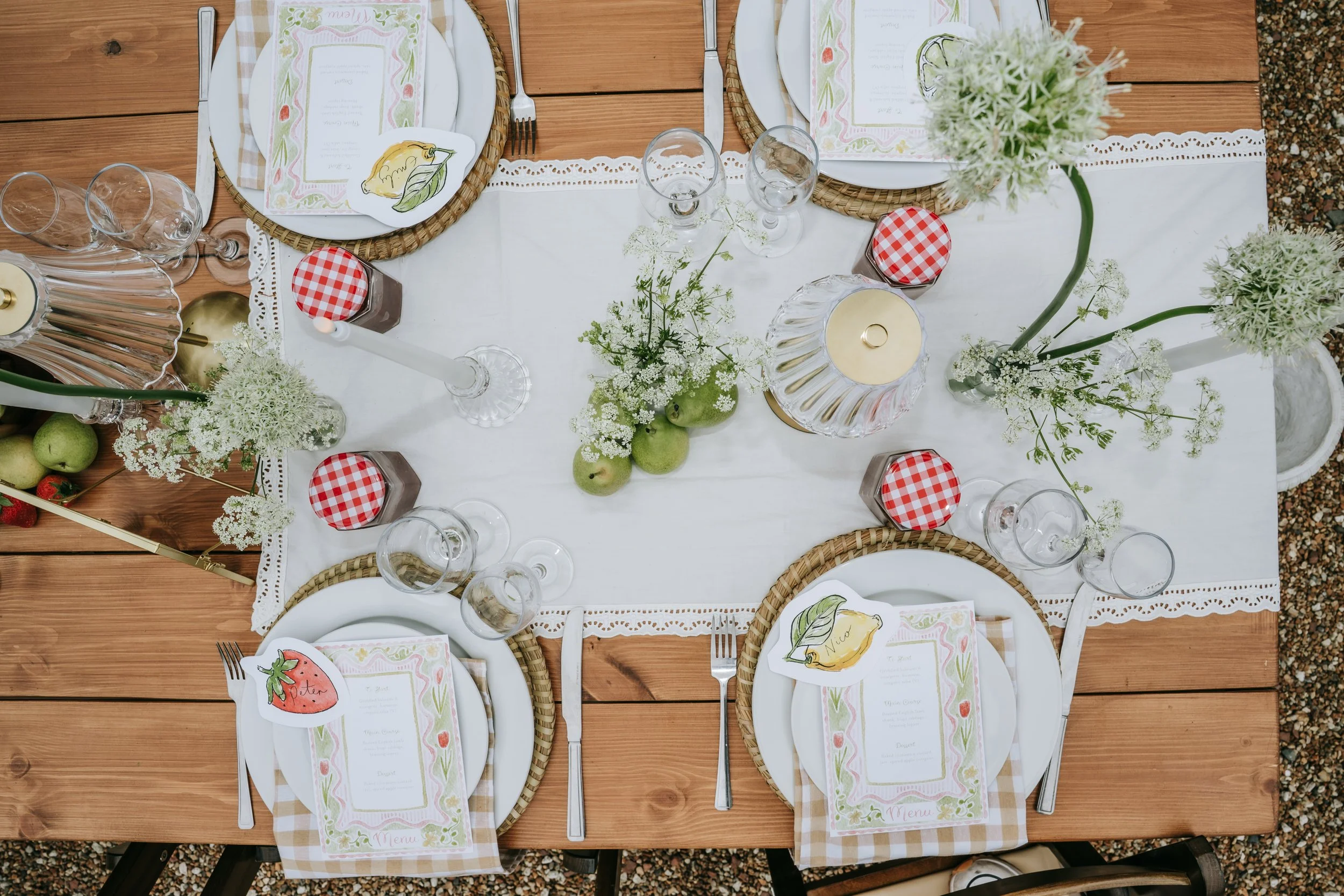 Top-down view of a table set for a meal with white plates, silverware, wine glasses, and decorative flowers and fruits, on a wooden table with a white table runner.