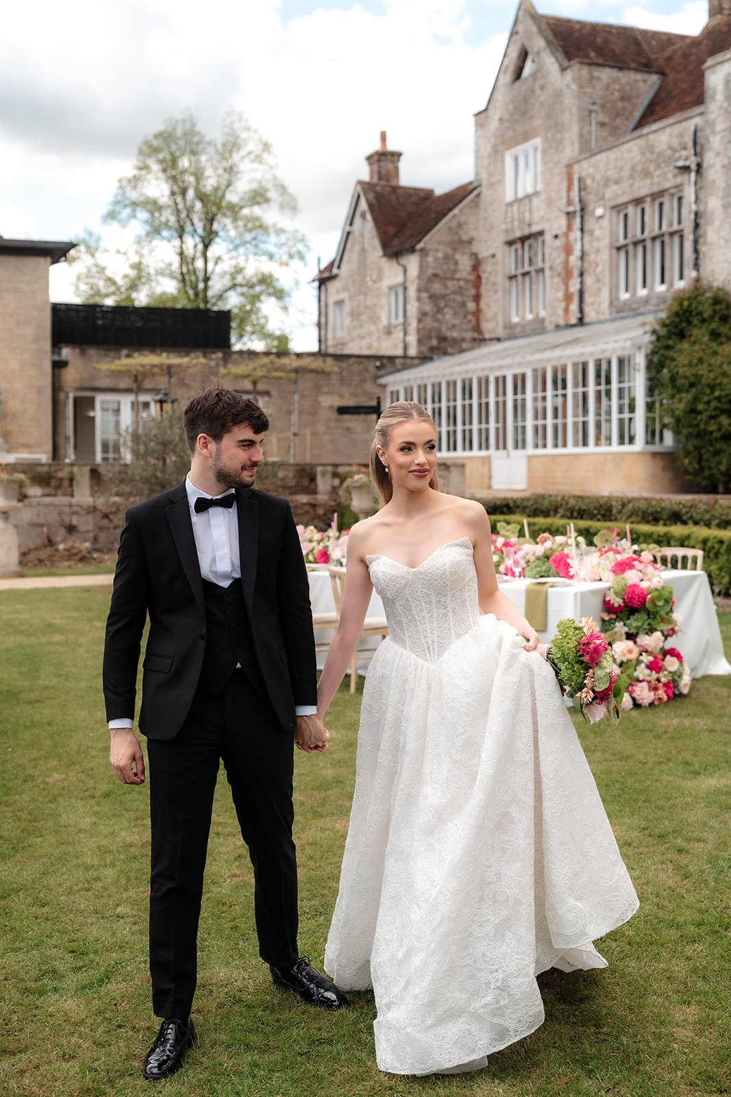A bride and groom holding hands, walking outside in a garden area with a table of pink and white flowers in the background, and an old building with large windows and ivy on the wall.