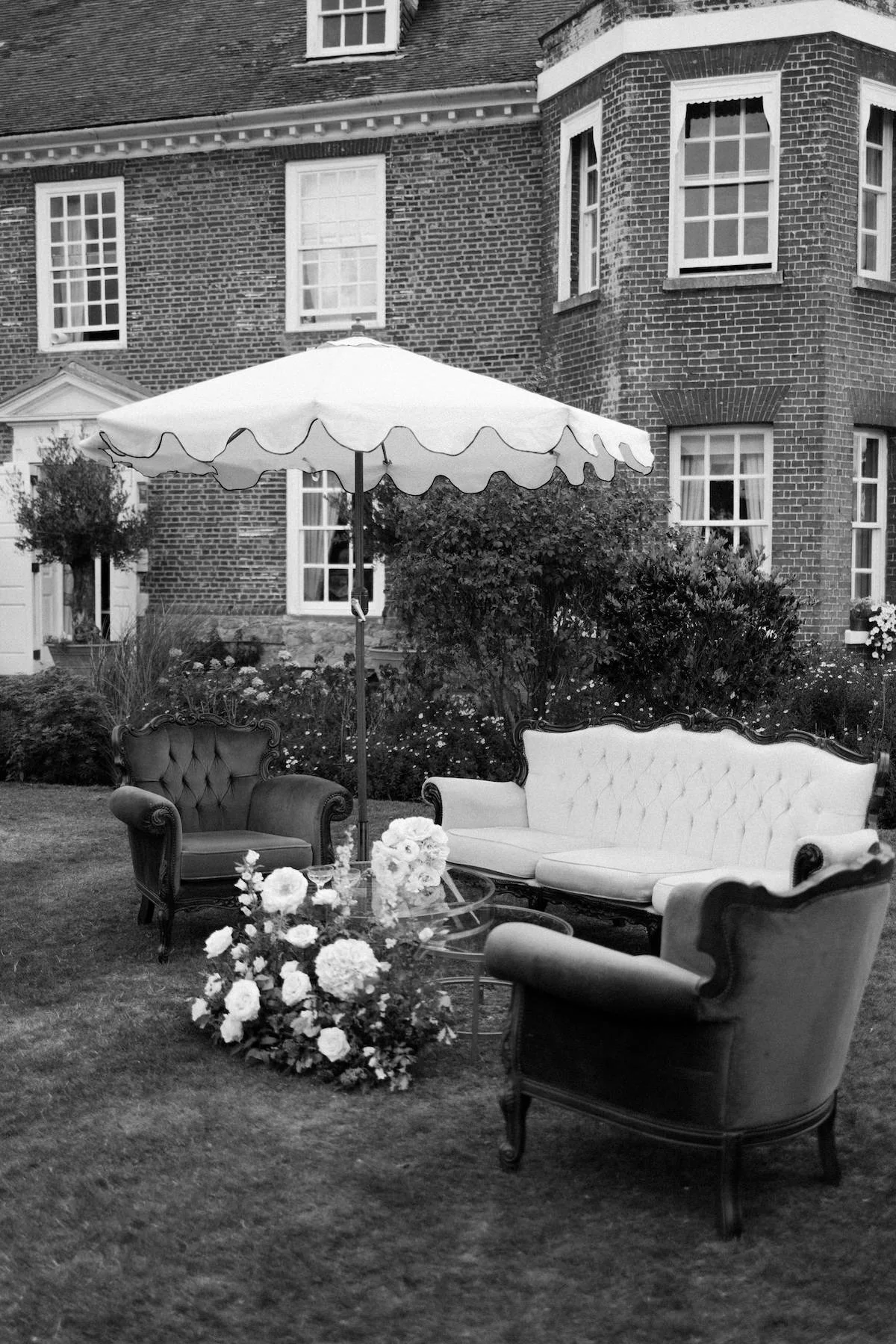A black and white photo of a backyard patio with antique-style furniture, including a sofa and armchair, around a small table with flowers, under a large umbrella. A brick house with multiple windows and a garden with bushes and flowers are in the ba