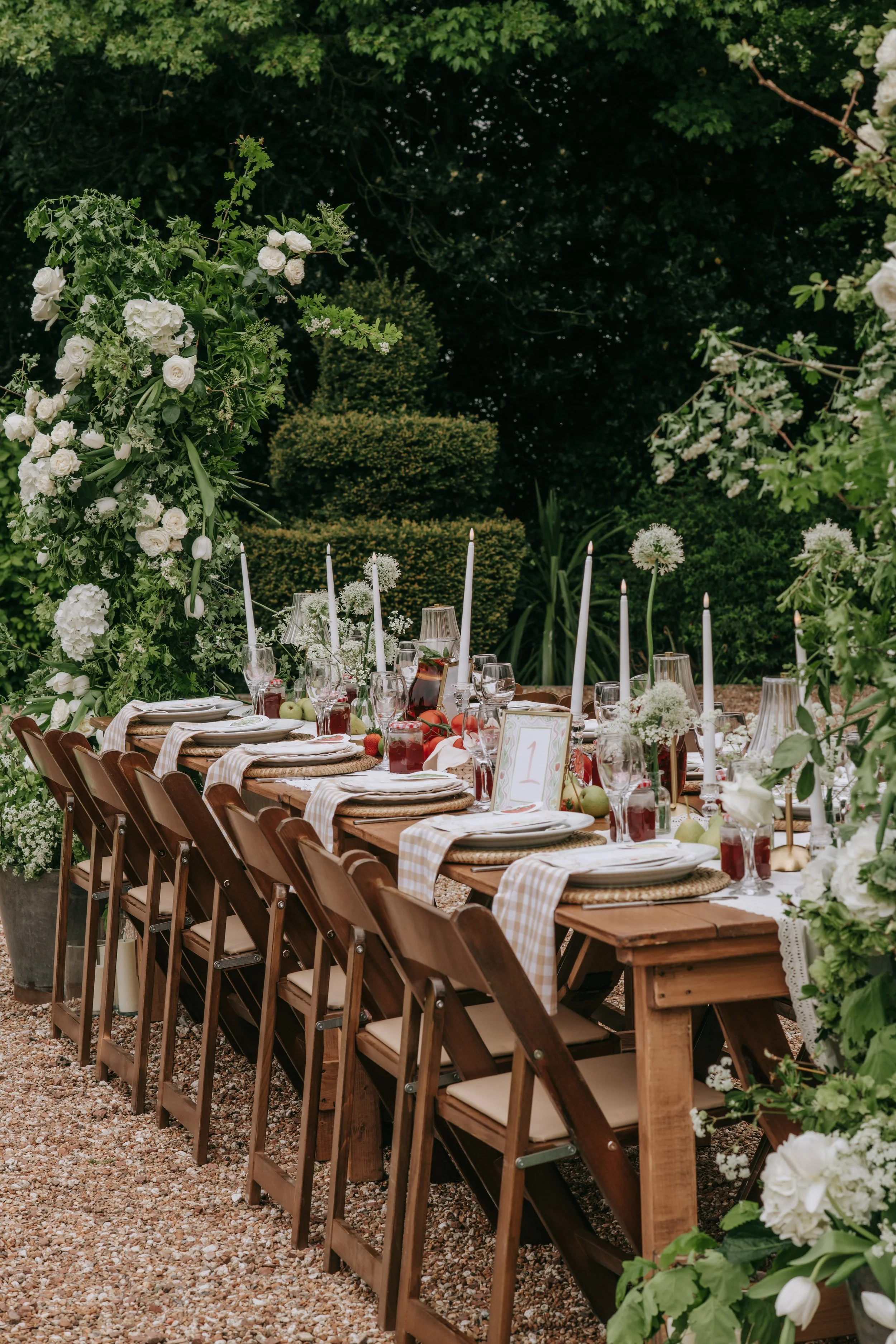 An outdoor wedding reception table decorated with white flowers, tall white candles, glasses, plates, and a table number, set amidst lush green foliage and bushes.
