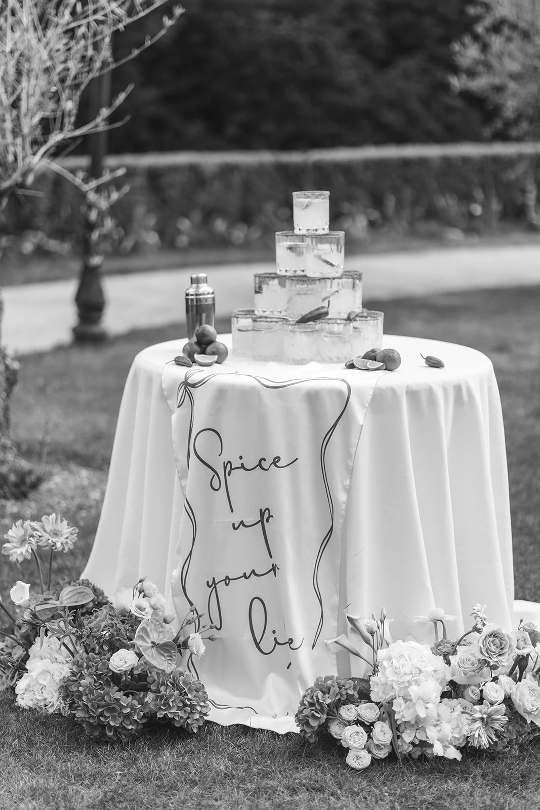 A table with a floral tablecloth reading 'spice up your life,' topped with a pyramid of ice blocks, fruits, and a shaker, surrounded by flowers and located outdoors.