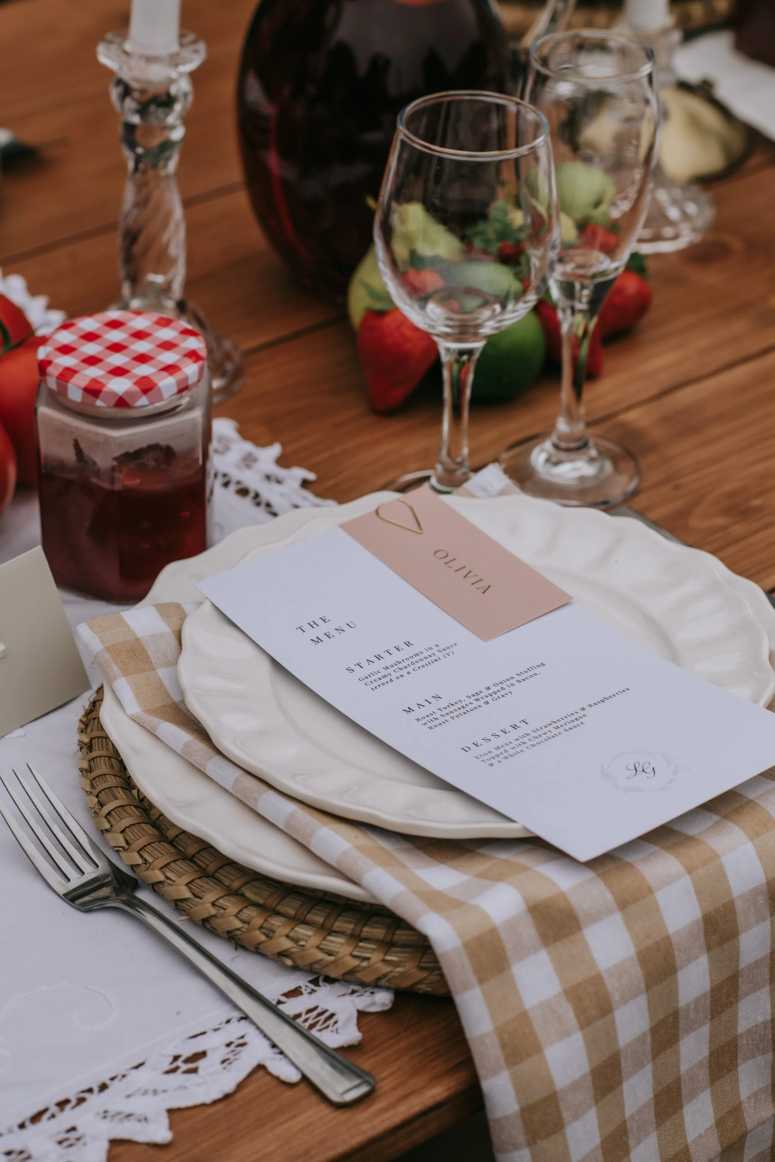 A table setting with a checked beige and white cloth napkin, a white plate with a menu on top, two empty wine glasses, a knife, and a fork on a wooden table. There are strawberries, a jar of jam with a red checkered lid, a dark bottle, and some fruit