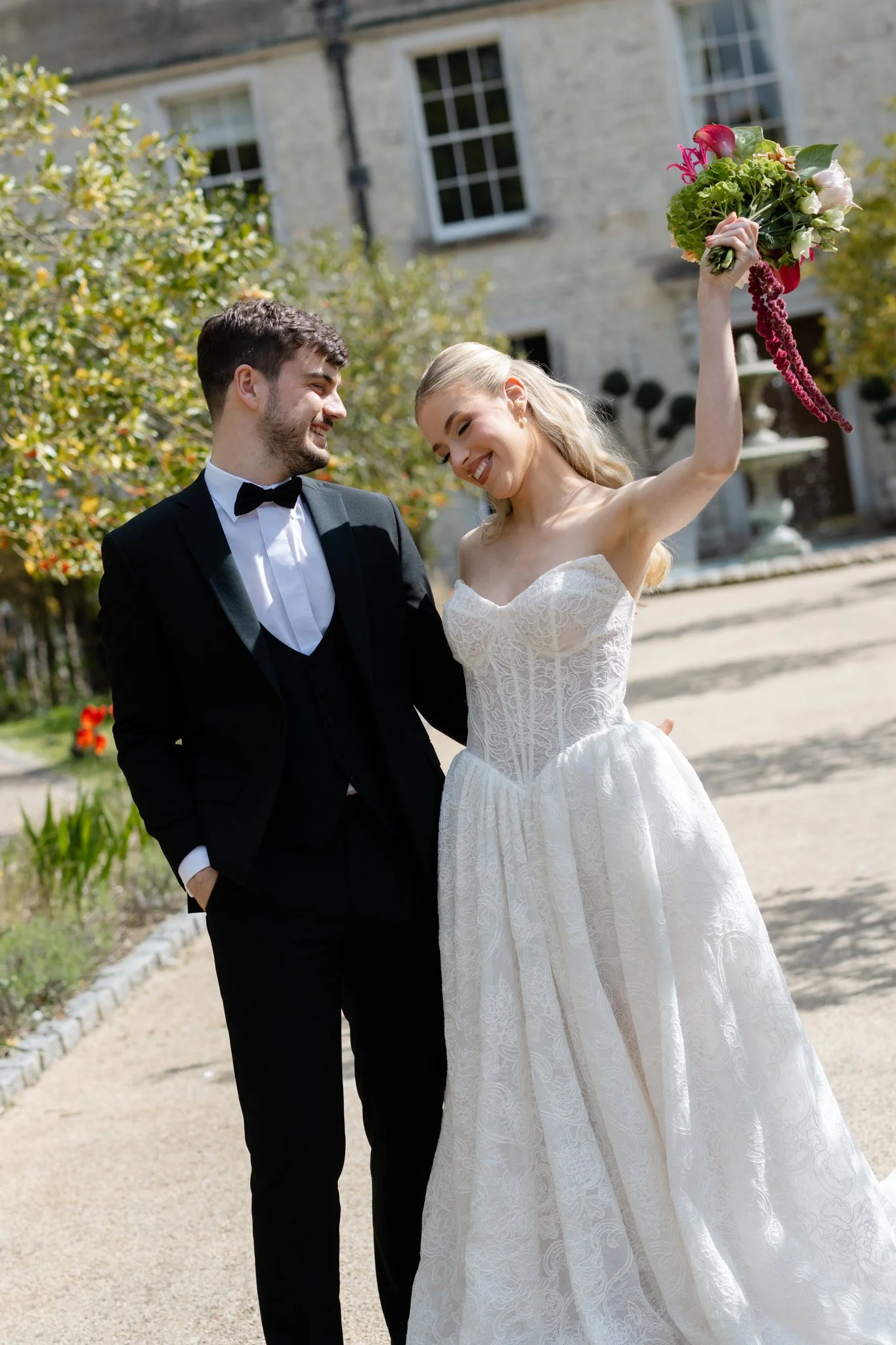A newlywed couple in wedding attire smiling and enjoying each other's company outdoors. The bride is holding a colorful bouquet of flowers high in the air. The groom is wearing a black tuxedo with a bow tie, and the bride is in a strapless white lace