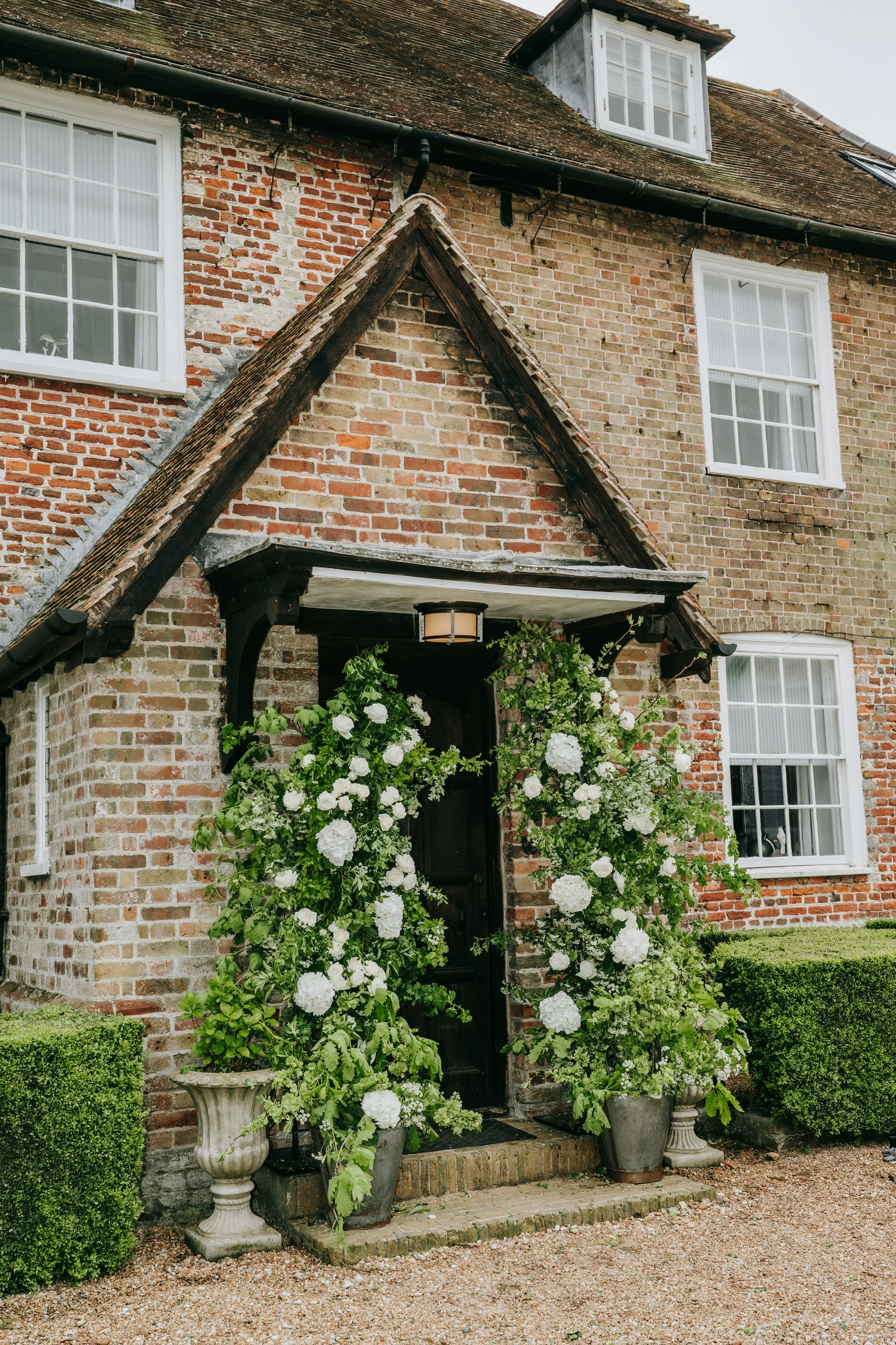 A brick house with white-framed windows and a dark front door surrounded by lush green plants and white flowers in vintage pots.