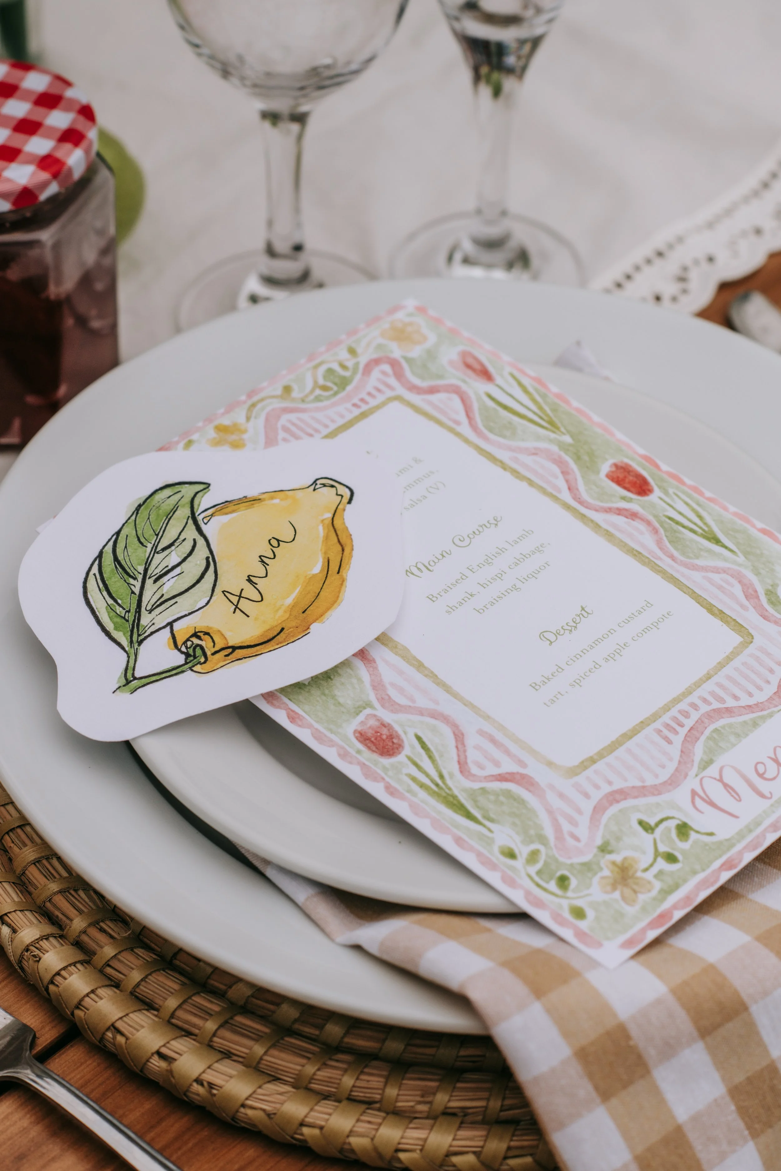 A place setting with a white plate, a menu with colorful floral border and a lion illustration, a woven placemat, a fork, and a glass on a wooden table with a checkered napkin.
