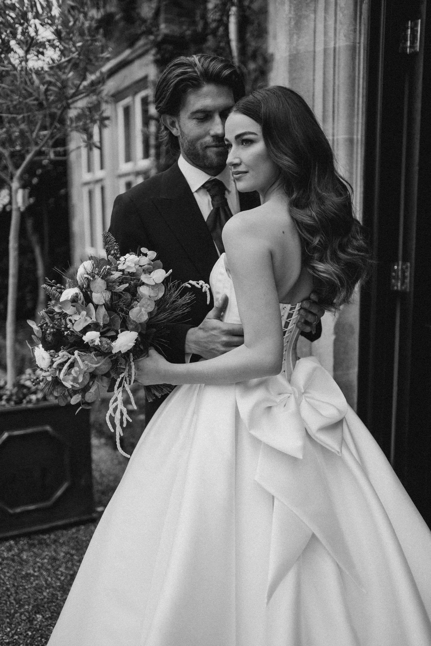 A black and white photo of a bride and groom standing close together outside. The groom has dark hair and wears a dark suit and tie, holding a bouquet of flowers. The bride has long wavy hair, wears a strapless wedding dress with a large bow at the w