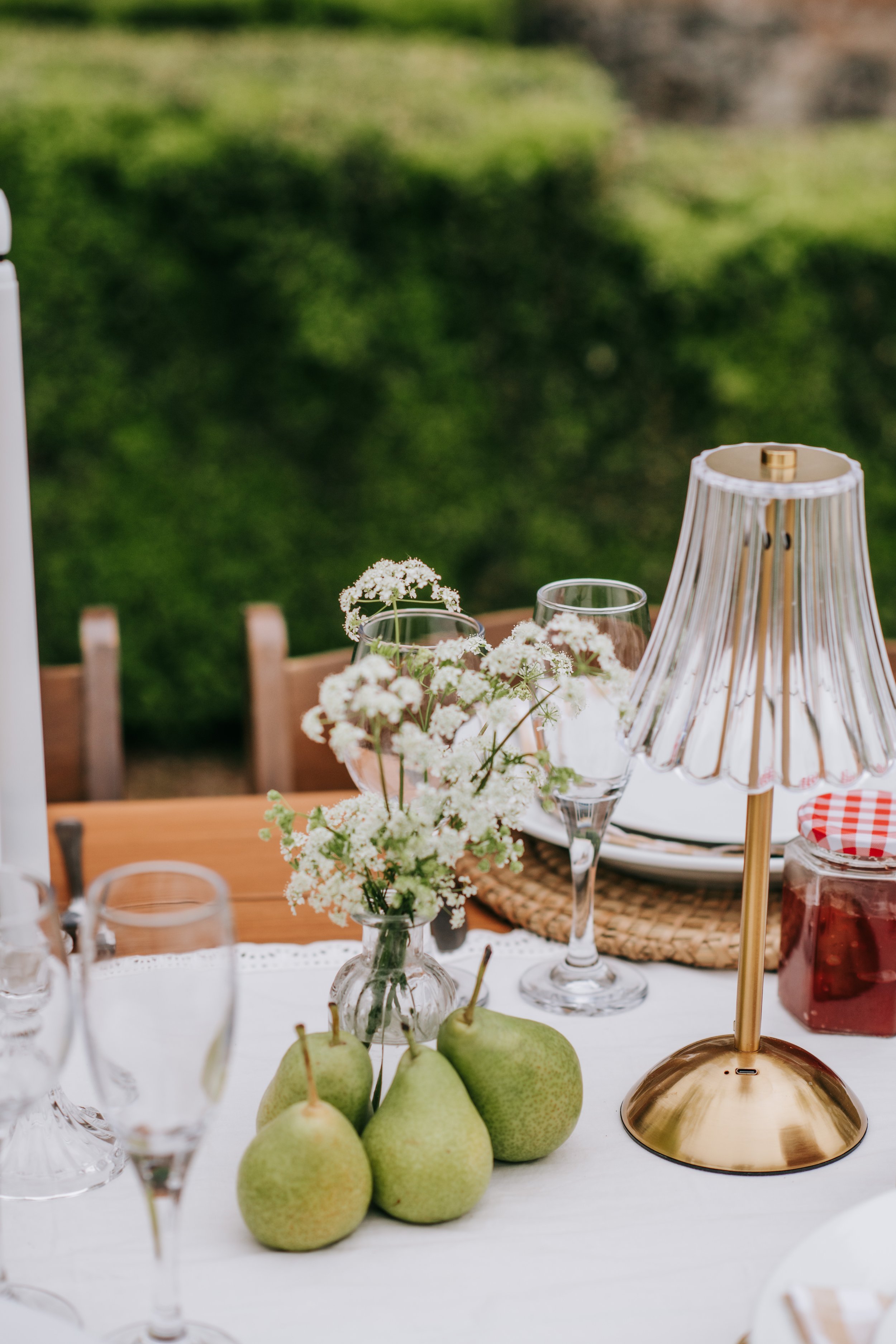A table set outdoors with a white tablecloth, decorated with small white flowers in a glass vase, green pears, empty wine glasses, a brass lampshade, and jars of jam in the background. Green foliage is visible in the blurred background.