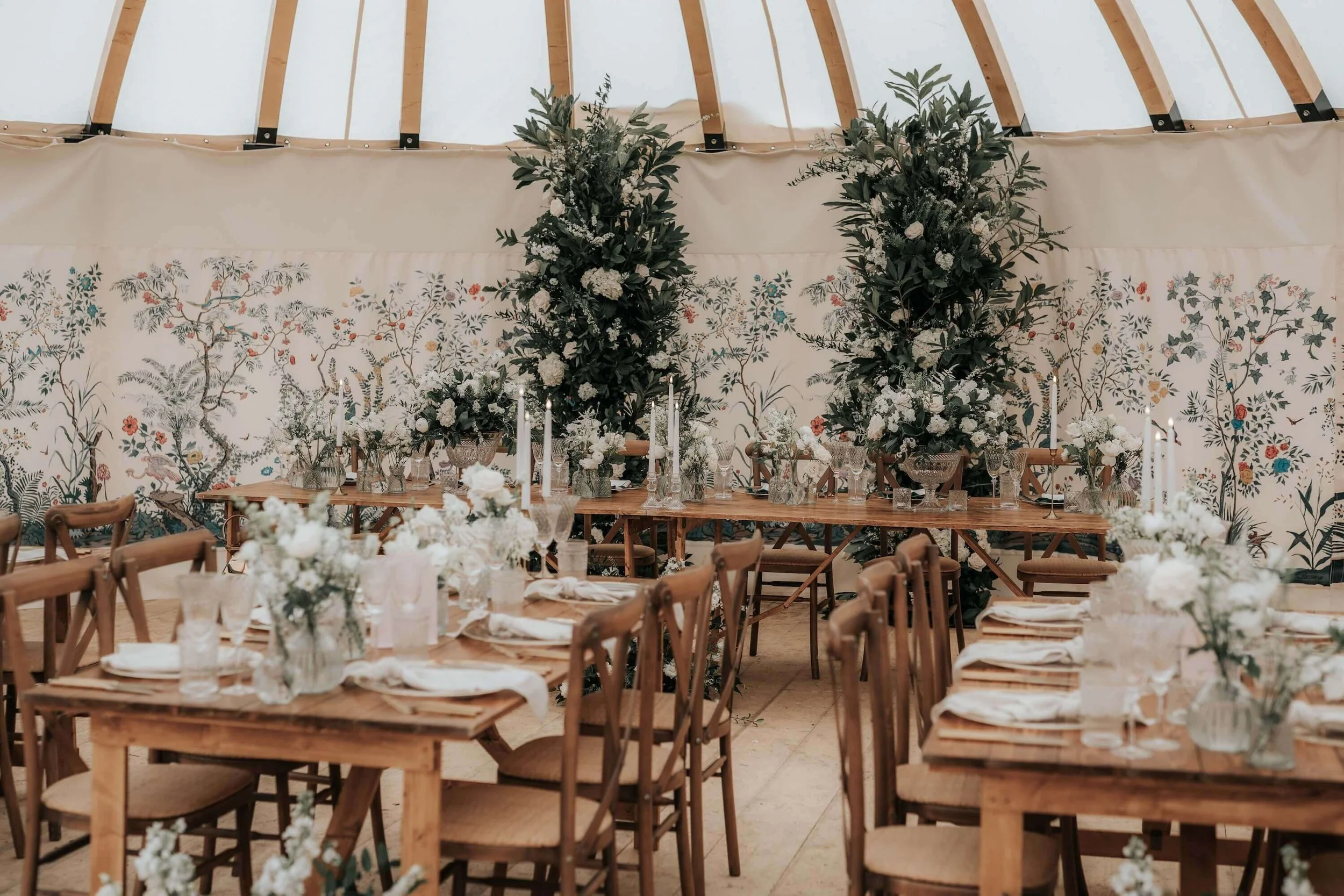 A decorated indoor event space with tables and chairs, floral centerpieces, and a backdrop of large greenery arrangements and painted wallpaper with floral patterns.