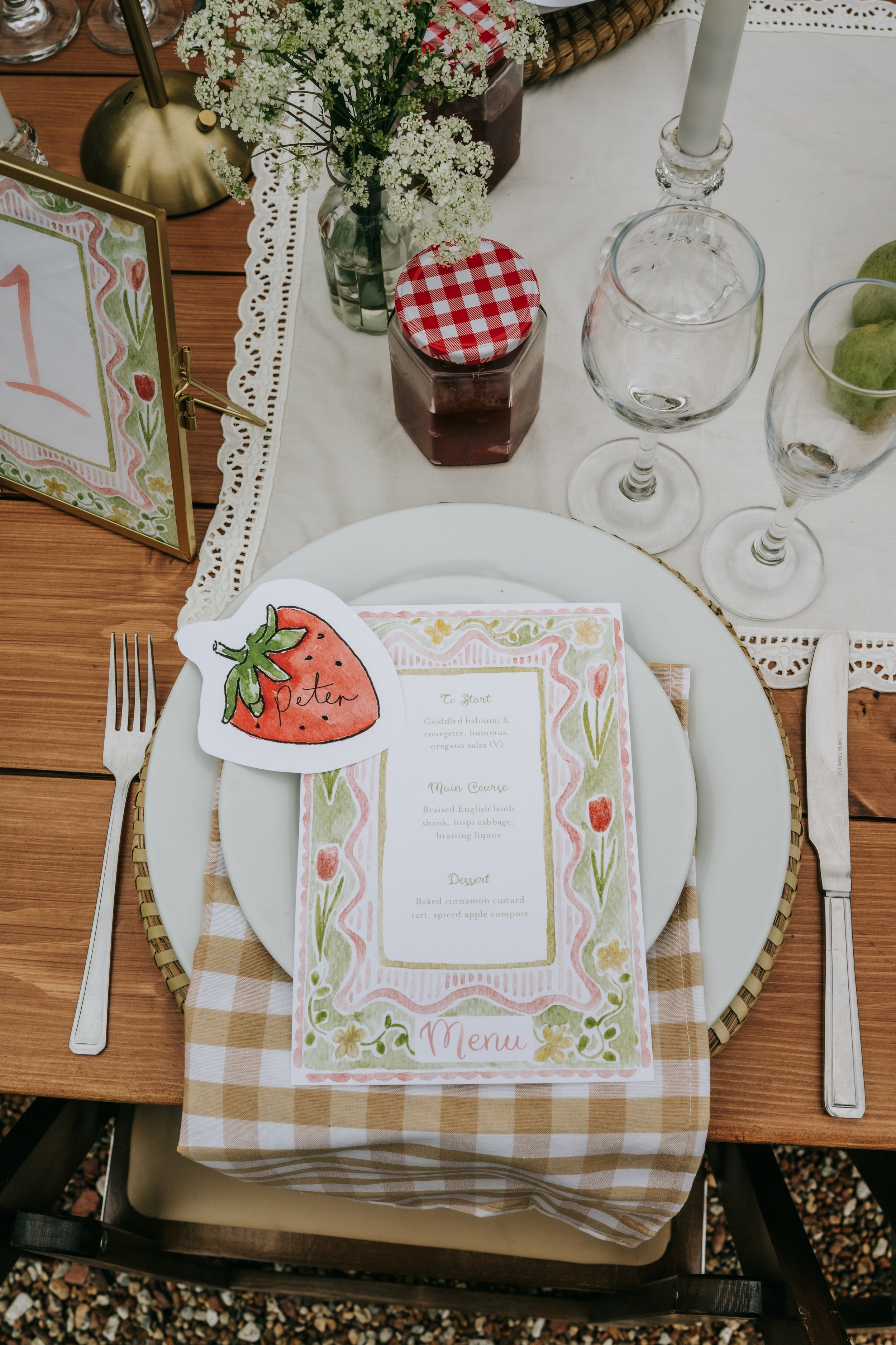 A table setting with a menu, a fork, a knife, a water glass, a wine glass, a candle, a small jar of jam with a red checkered lid, a flower bouquet in a glass vase, and a strawberry place card reading 'Peter'. The table has a checked tablecloth and a 