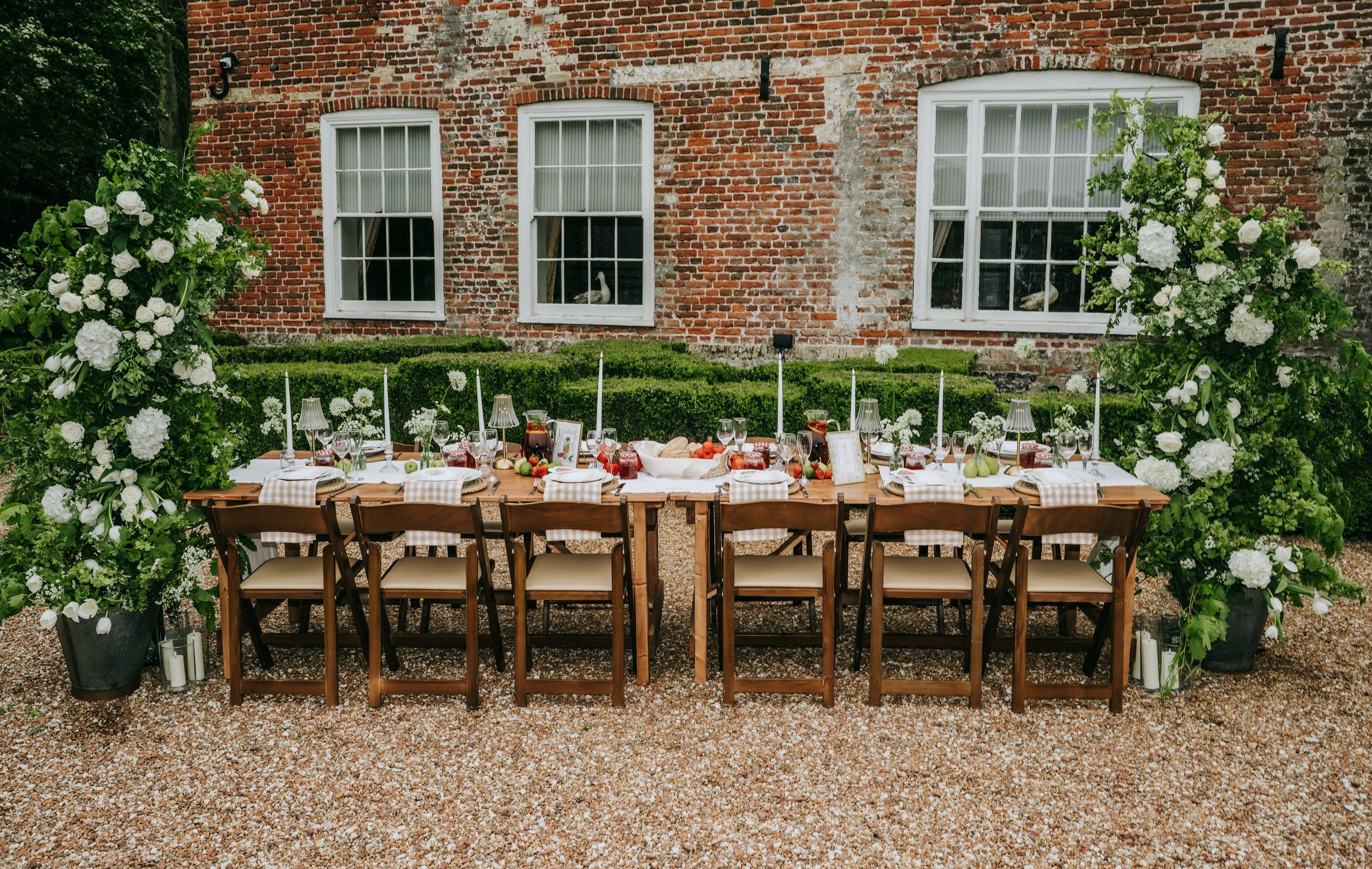 A long wooden table set for an outdoor celebration with white flowers, candles, and tableware, located in front of a brick building with three large windows with white frames, flanked by tall floral arrangements.
