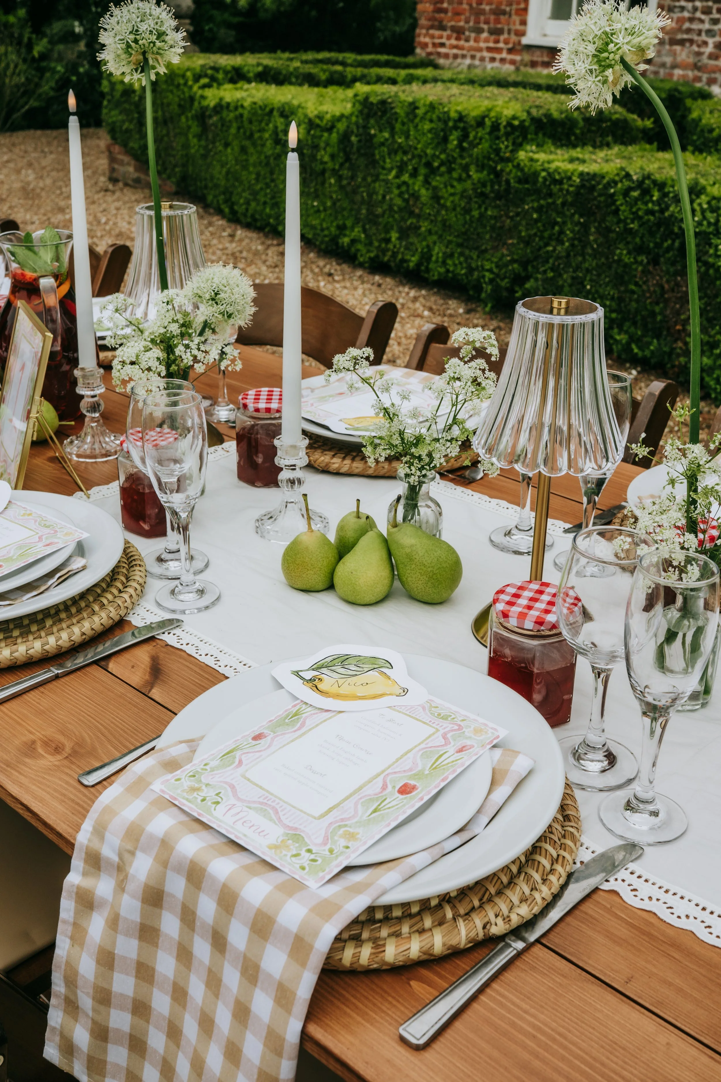 Outdoor dining table set with white plates, beige checkered napkins, glassware, floral centerpieces, candlesticks, decorative lamps, and fresh pears, arranged in a garden setting with bushes and a brick house in the background.