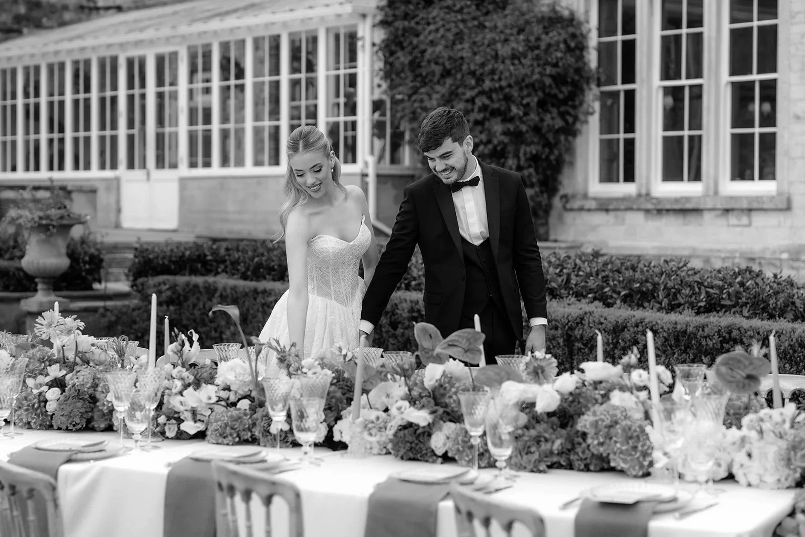 Black and white photo of a bride and groom standing outside next to a decorated table, smiling and looking down at the table.
