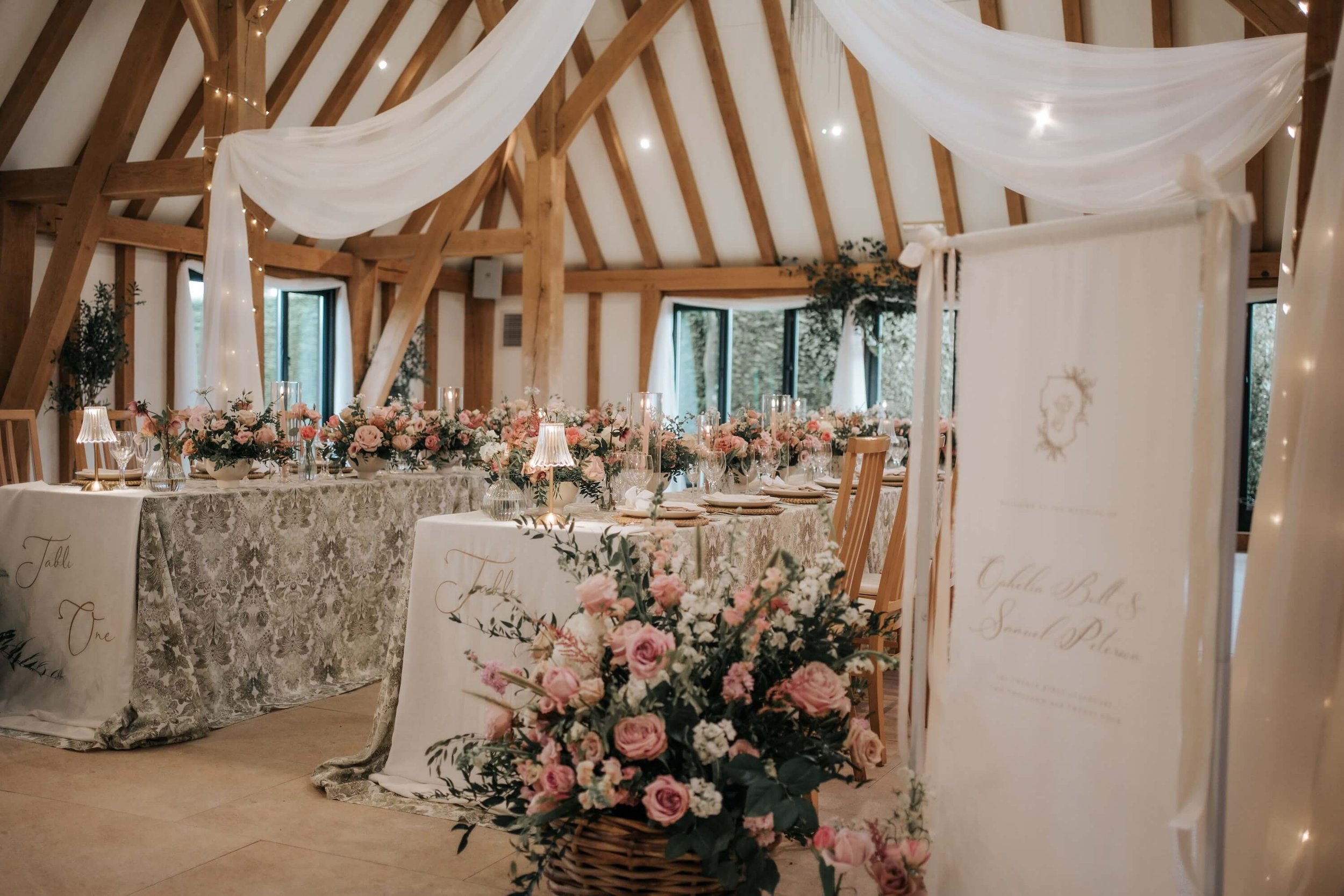 Wedding reception setup with a long table decorated with pink and white flowers, candles, and lace tablecloths inside a wooden banquet hall with draped white fabric and string lights overhead.