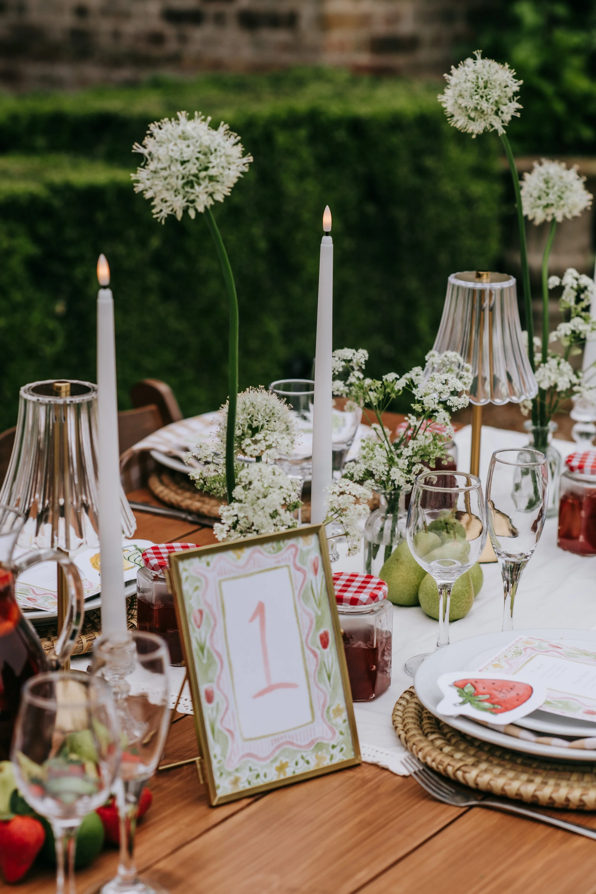 A decorated outdoor table set for a celebration with white flowers, candles, a table number card, and glassware.