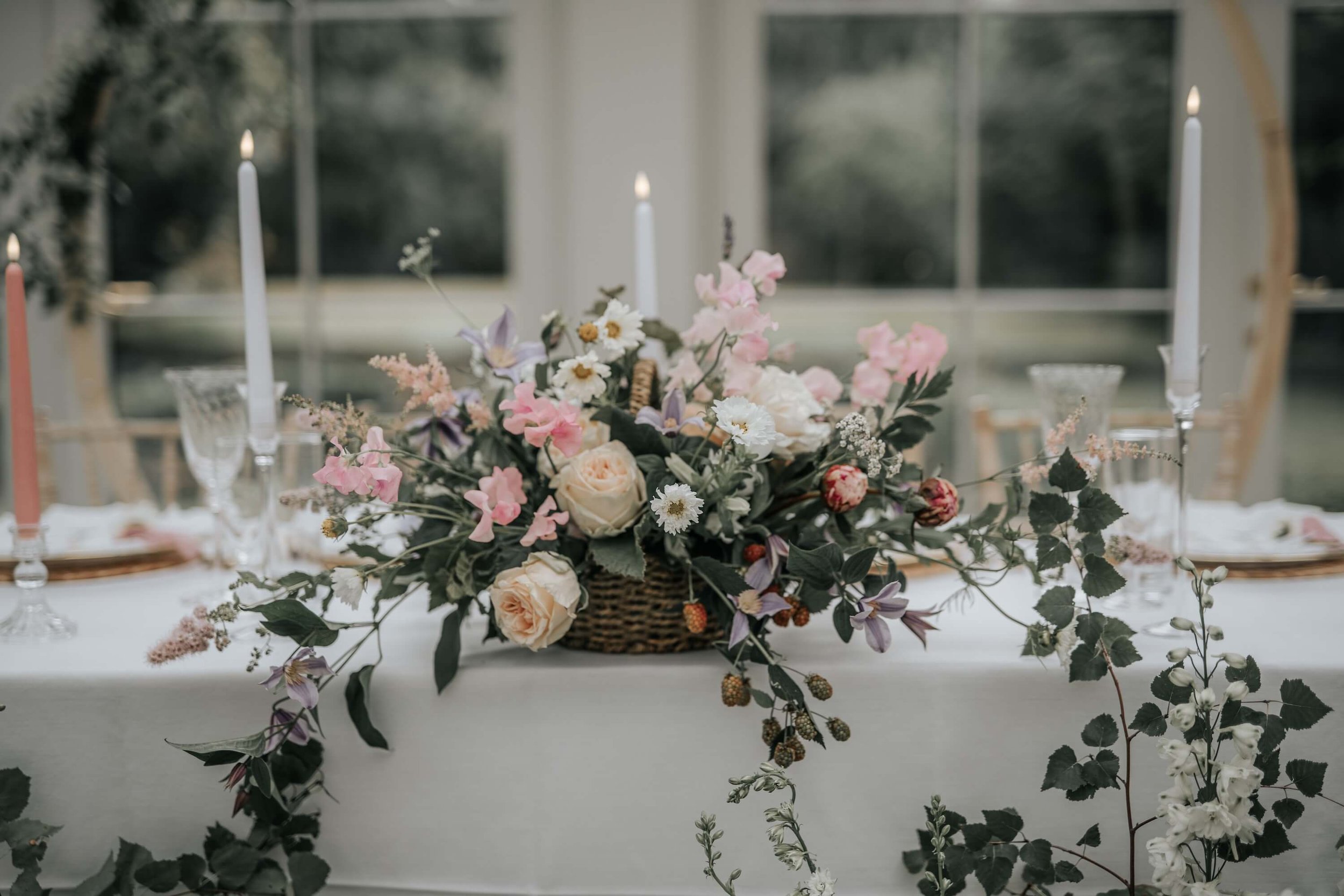 A flower arrangement with pink, white, and purple flowers in a wicker basket on a table with a white tablecloth, decorated with pink candles and glassware, set in a bright room with large windows in the background.