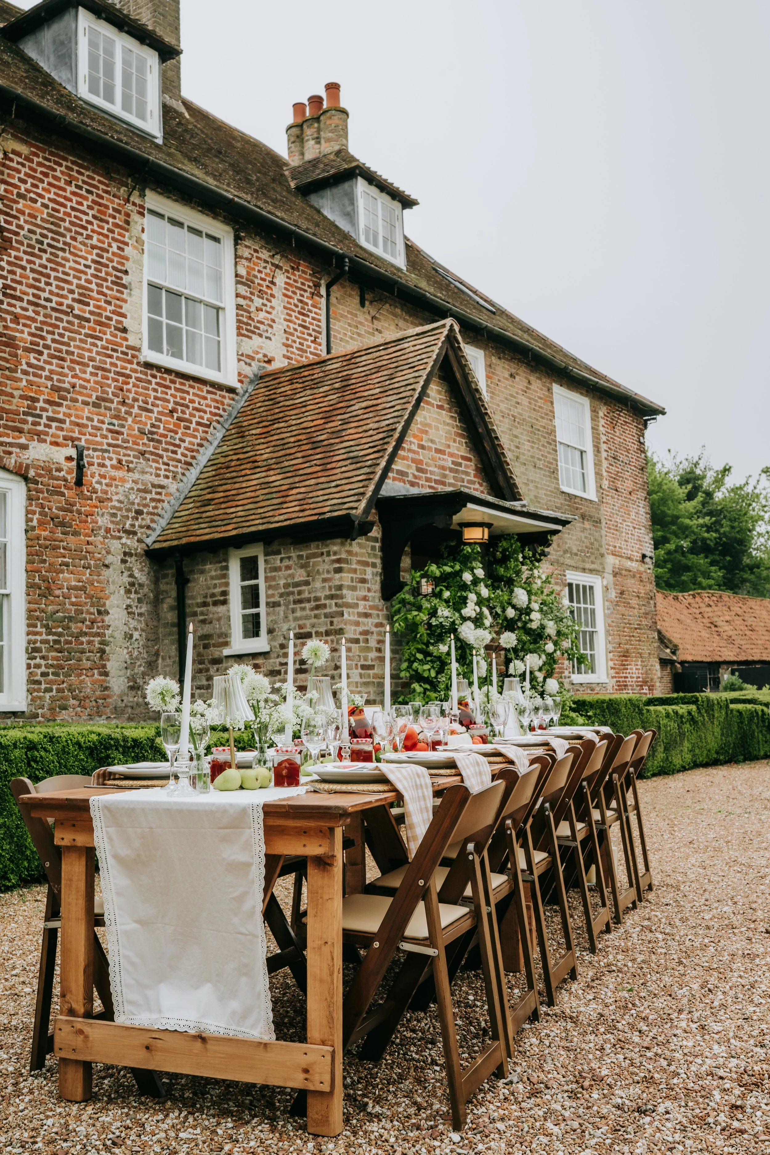 An outdoor banquet table set up in front of a historic brick house with a landscaped lawn and trees in the background, decorated with white flowers, candles, and tableware.