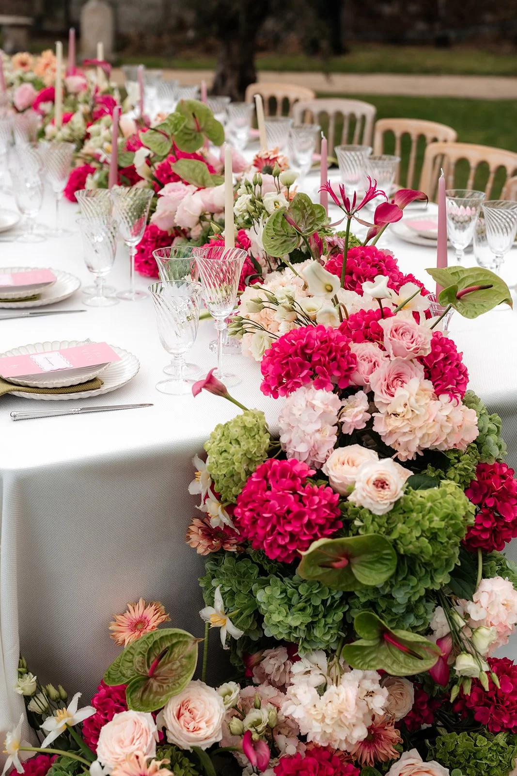 Elegant outdoor dining table decorated with colorful pink, white, and green floral arrangements, surrounded by white chairs and a white tablecloth.