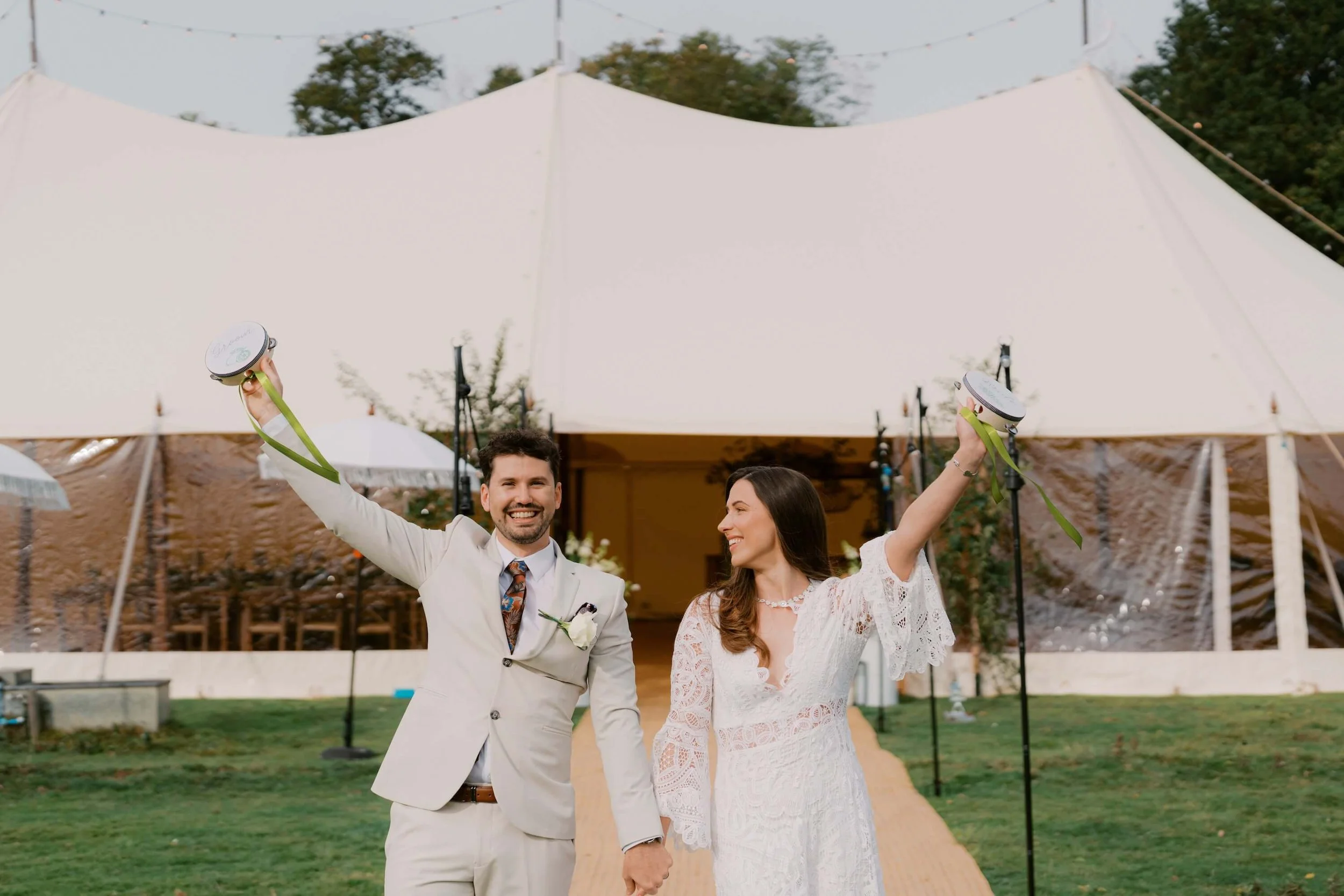 A newlywed couple celebrating outdoors, holding their wedding bells and smiling. The man is wearing a white suit with a floral tie, and the woman is in a white lace wedding dress. They are standing on a pathway in front of a large white tent with string lights, trees, and a grass lawn in the background.