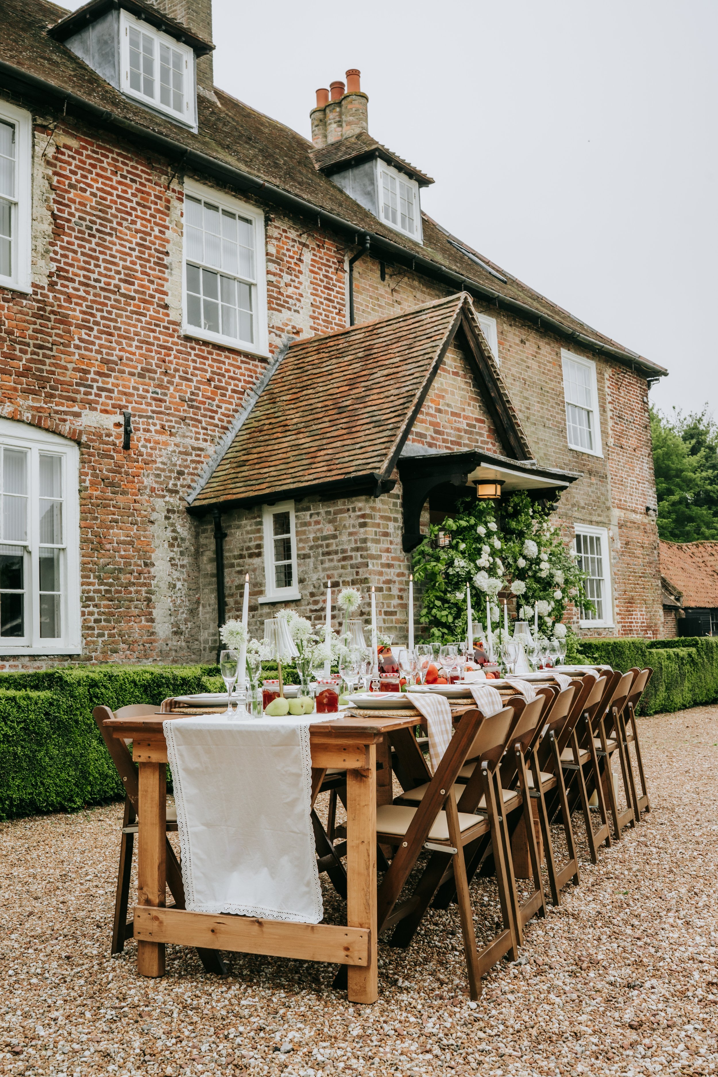 Long outdoor dining table set for a meal, with wine glasses, candles, and floral decorations, placed on a gravel yard in front of a historic brick house with multiple windows and a sloped roof.