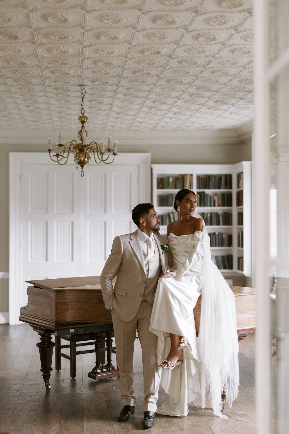 A bride in an off-the-shoulder white wedding dress sitting on a piano with a groom in a beige suit standing beside her in a room with a vintage ceiling, chandelier, and bookshelves.
